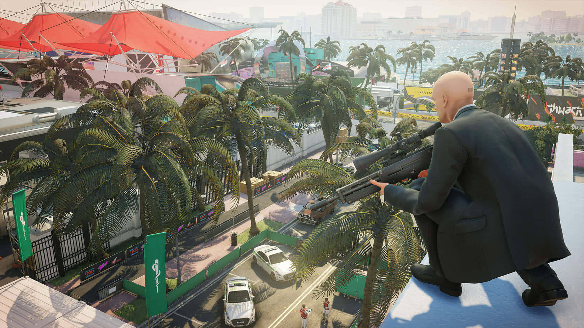 留下你的標記 — 立即體驗備受好評的《最佳 Hitman 2》 壁紙 — 一個光頭穿西裝的男人蹲在屋頂上，用狙擊步槍瞄準棕櫚樹對面繁忙的熱帶城市景觀。