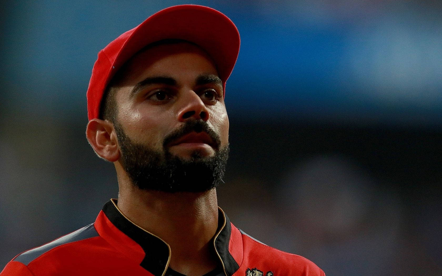 virat kohli rcb 桌布 壁紙 — 留鬚嘅板球球員近距離肖像，戴住紅色帽同波衫，喺模糊嘅球場背景下專注向前望。