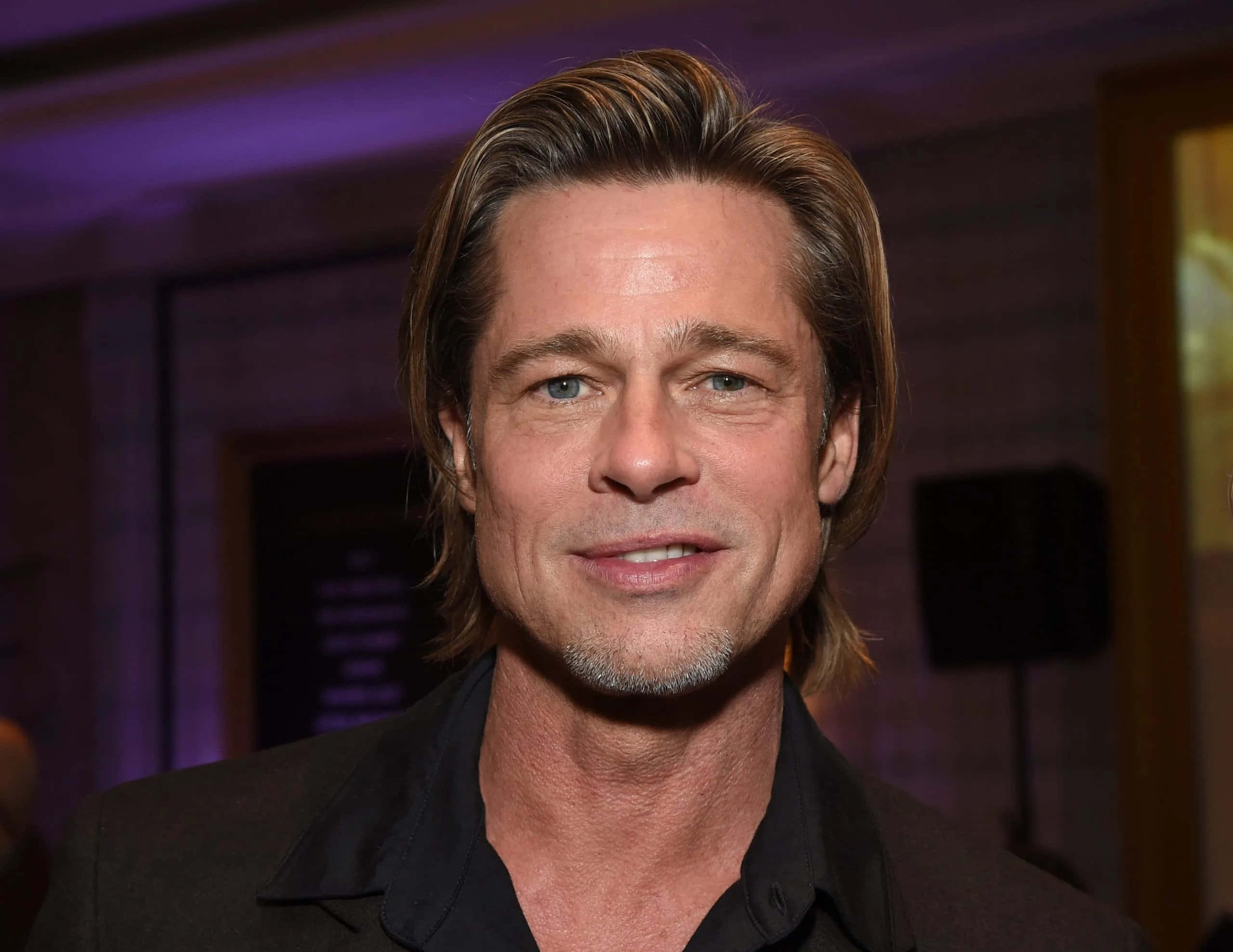 Brad Pitt 桌布 壁紙 — 畢彼特在室內活動中穿著深色西裝，在紫色環境燈光下溫暖地微笑。