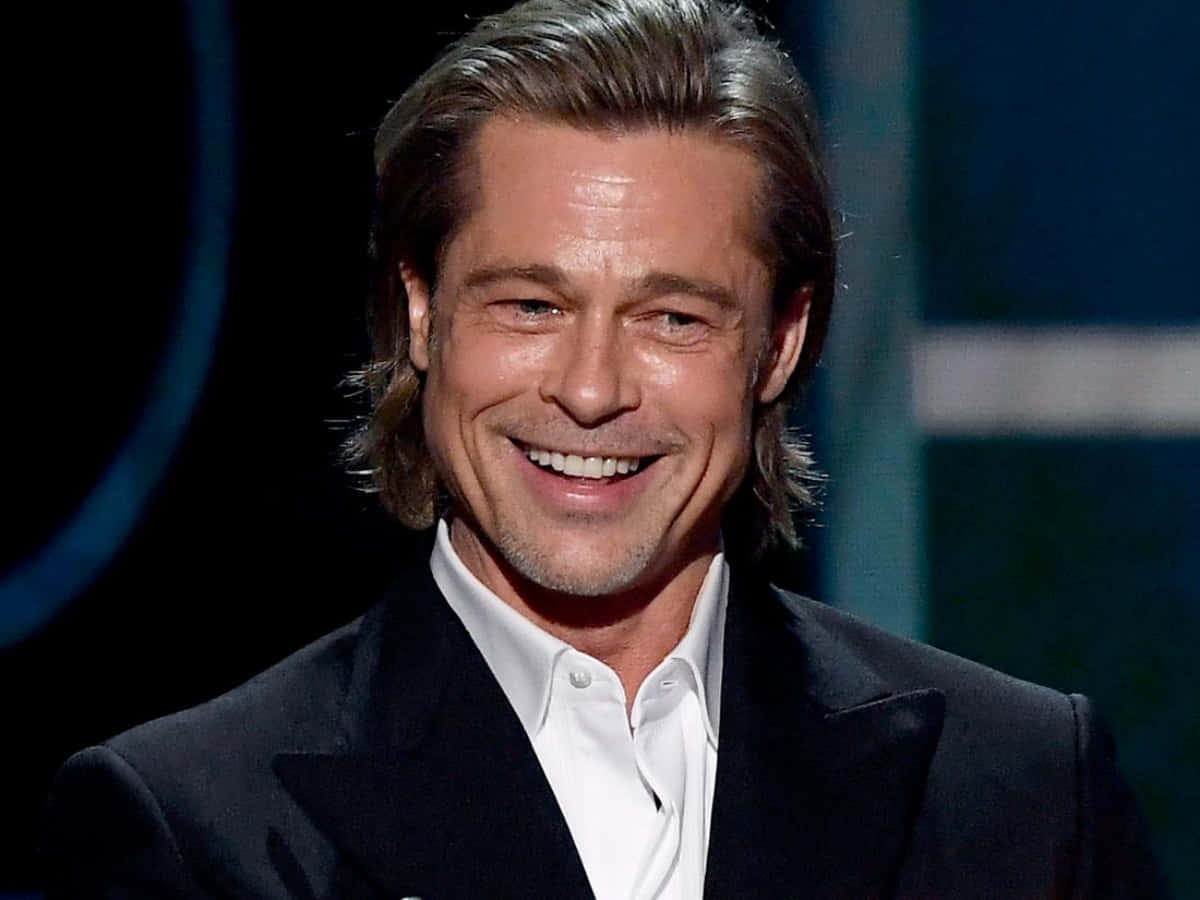 Brad Pitt 桌布 壁紙 — 畢彼特在舞台燈光下穿著黑色西裝和白色襯衫溫暖地微笑。