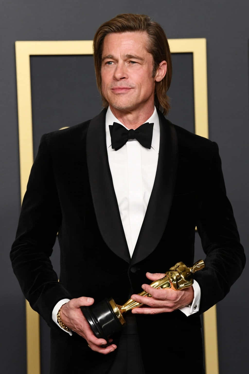 Brad Pitt 桌布 壁紙 — 畢彼特身穿黑色天鵝絨燕尾服，手持金色奧斯卡獎座，面帶含蓄微笑，背景為深灰色並配以金色邊框。