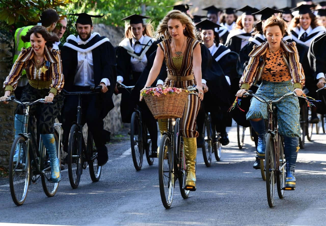 Mamma Mia 桌布 壁紙 — 畢業生戴著四方帽、穿著畢業袍，在陽光明媚的街道上騎單車，身穿色彩繽紛的圖案服裝，帶著花籃。