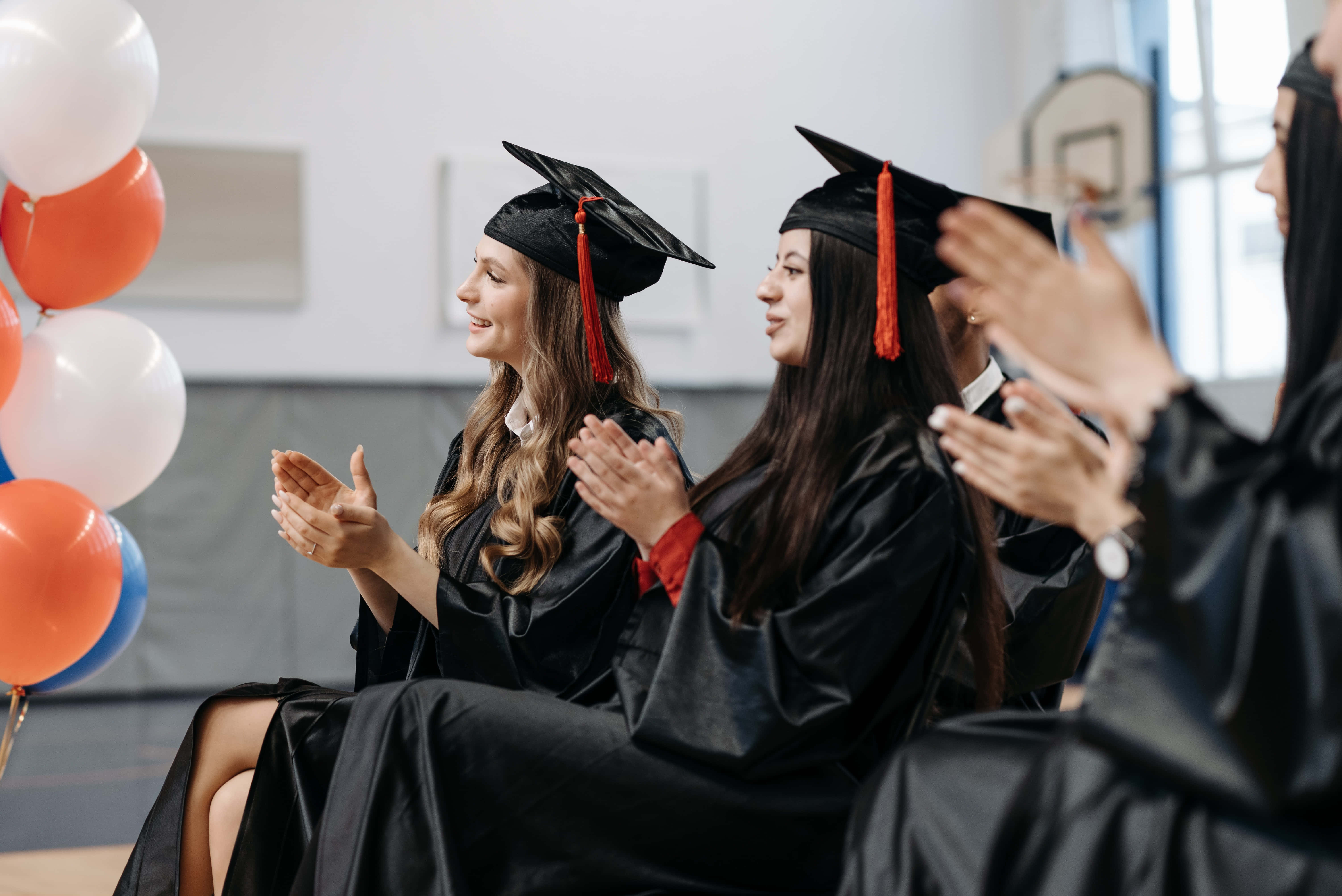 Graduation Ceremony 桌布 壁紙 — 畢業生身穿黑色四方帽和畢業袍，在明亮的體育館內，於色彩繽紛的氣球旁開心地鼓掌。