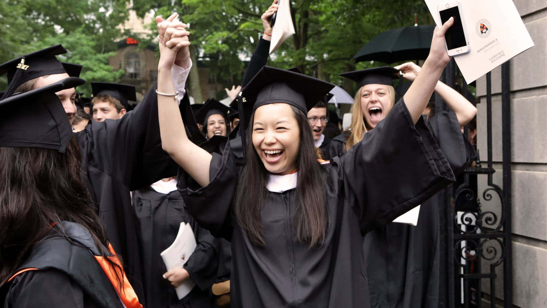 Graduation Ceremony 桌布 壁紙 — 畢業生身穿黑色學士帽和袍，在戶外綠樹下高舉文憑和手機，興高采烈地歡呼。