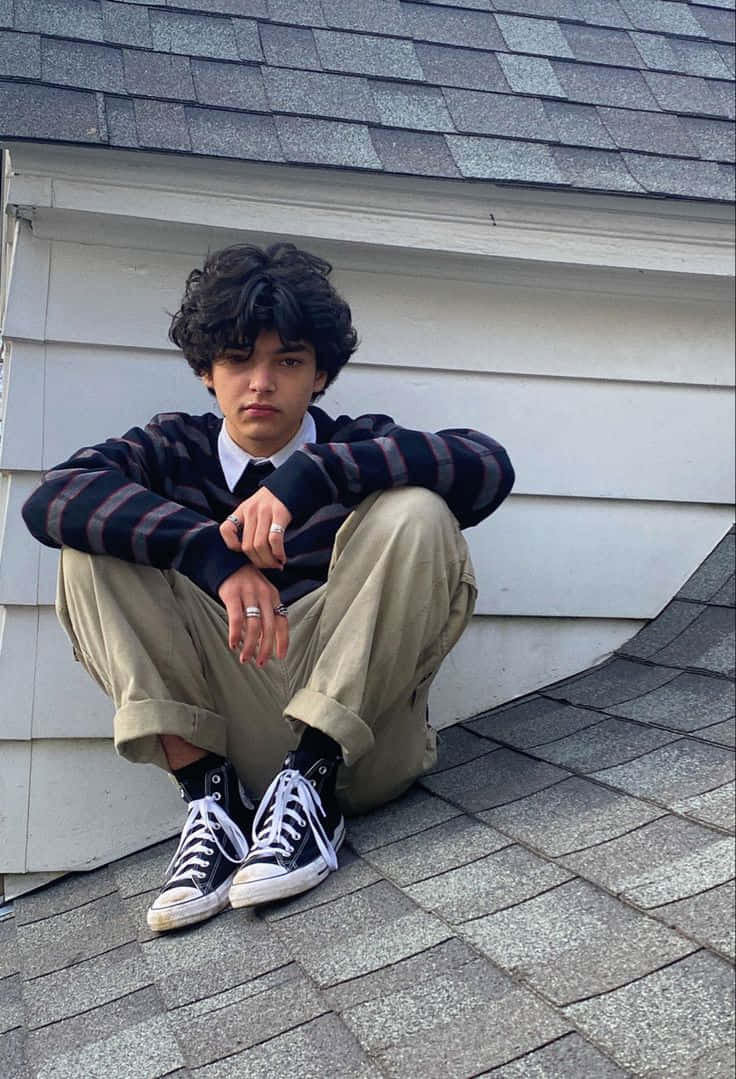 當代時尚男孩 壁紙 — 一個有捲髮的年輕人盤腿坐在灰色的瓦片屋頂上，穿著條紋毛衣和黑色運動鞋。