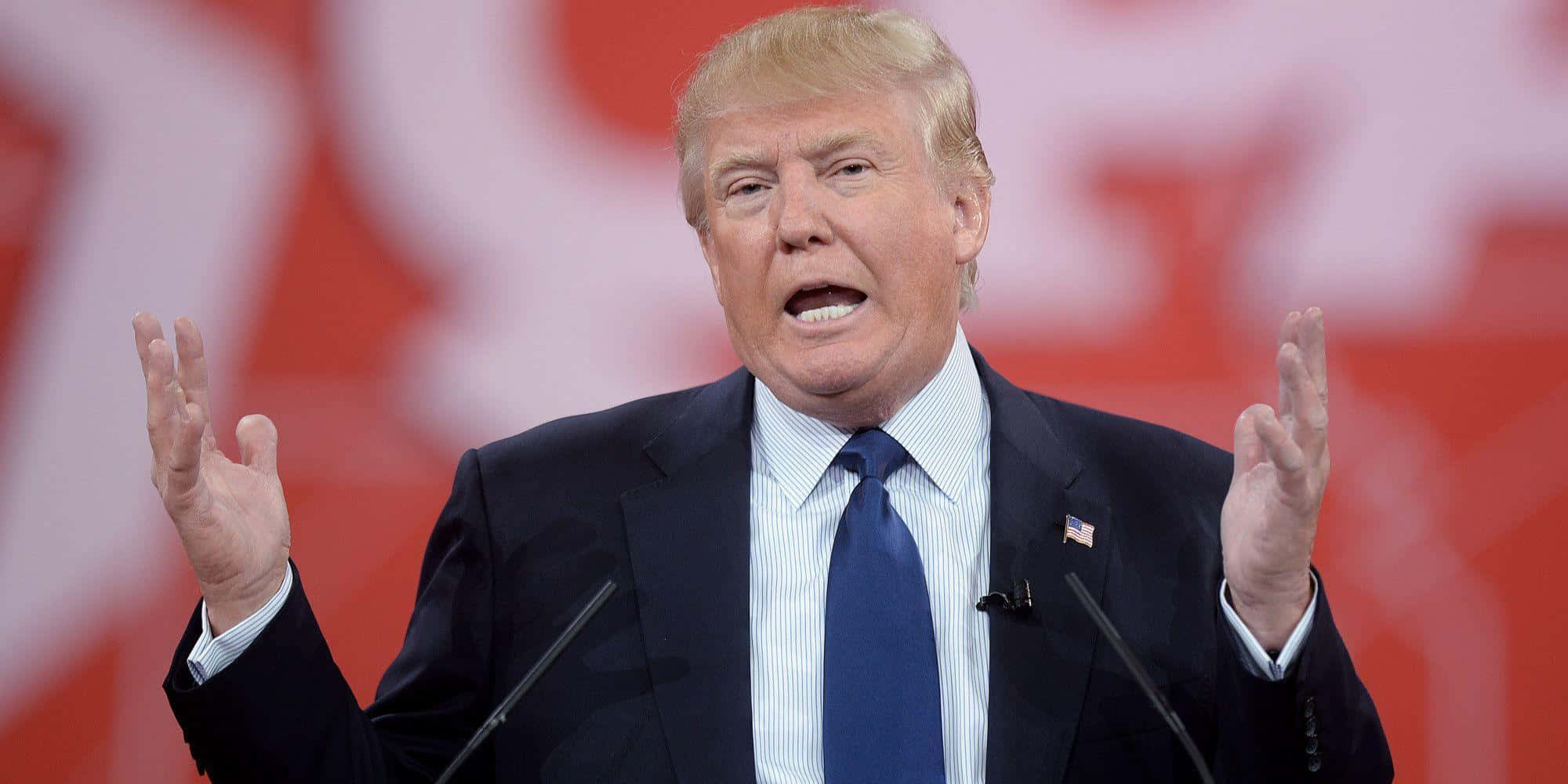 trump 桌布 壁紙 — 當勞·特朗普在紅色背景配白色字體的講台上，以強調的手勢發表演說。