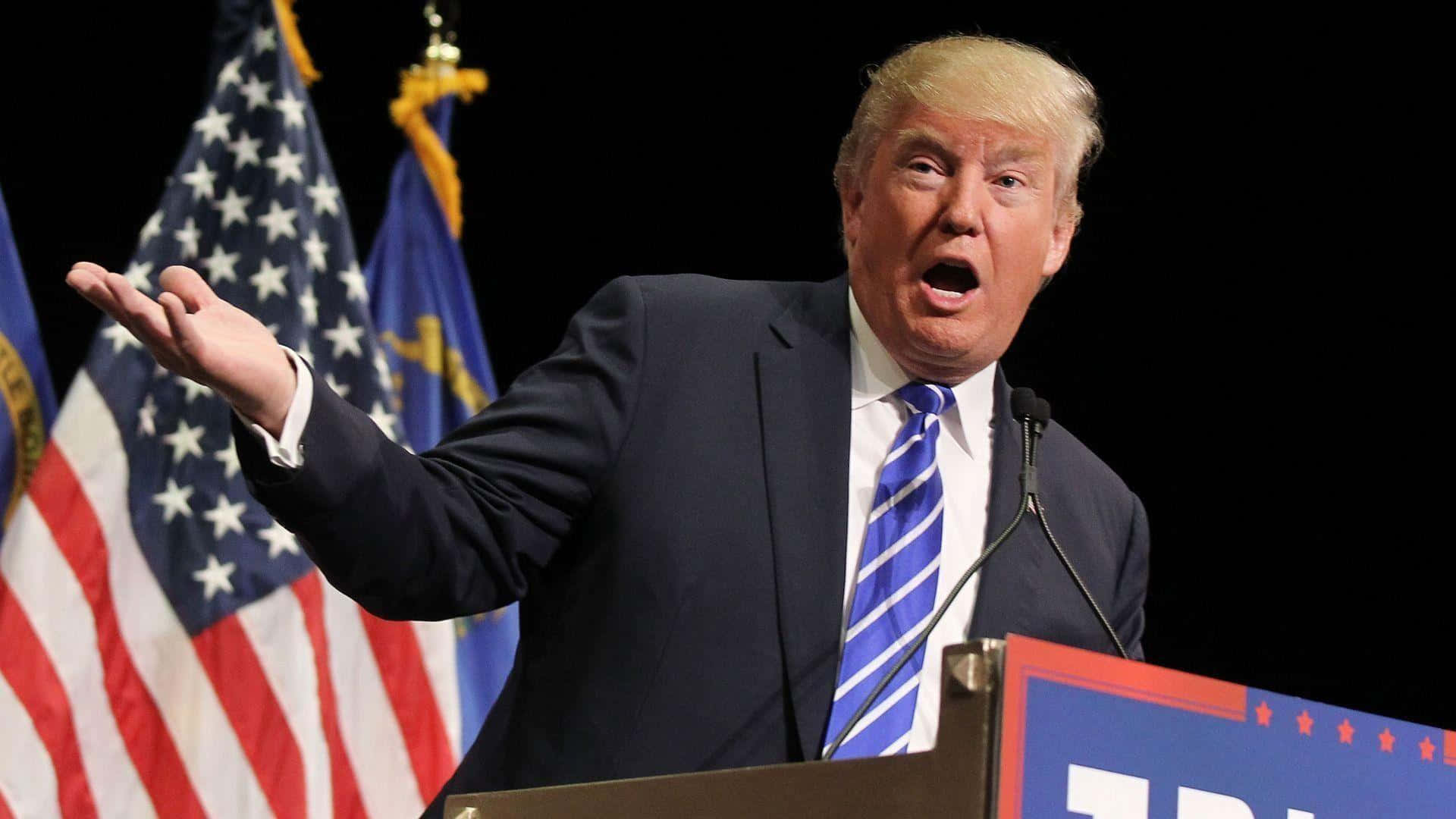 trump 桌布 壁紙 — 當勞·特朗普在講台上強調地做手勢，身後有美國國旗和州旗在舞台燈光下。