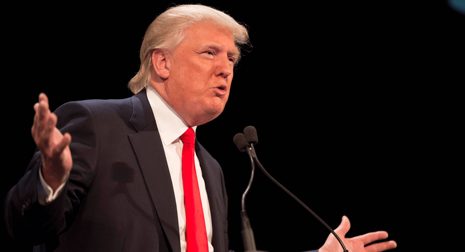 donald trump 桌布 壁紙 — 當勞·特朗普在黑色背景的明亮舞台燈光下，在講台上說話時強調地做手勢。