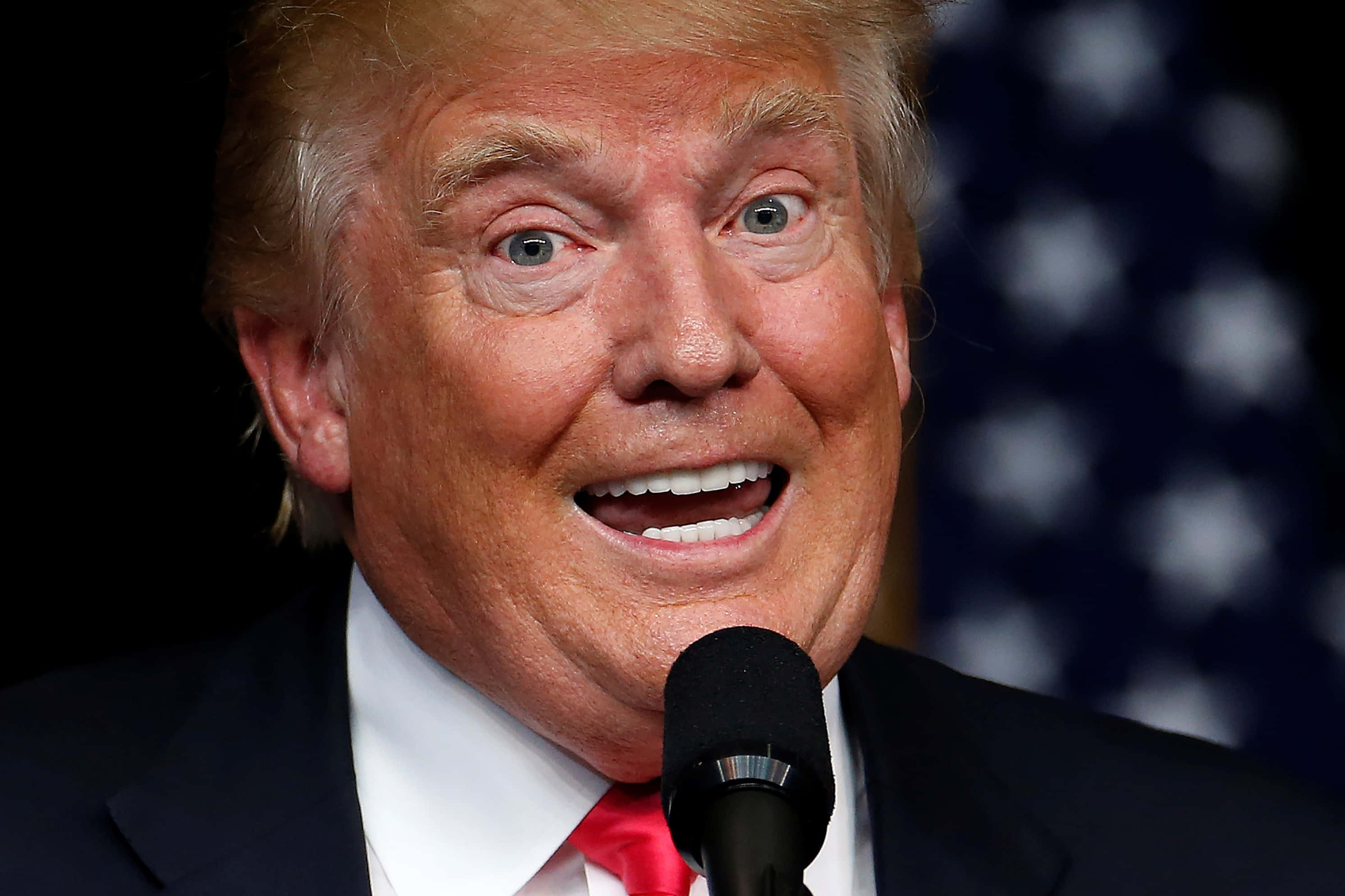 trump 桌布 壁紙 — 當勞·特朗普對著咪高峰講話的特寫鏡頭，背後有模糊的美國國旗。