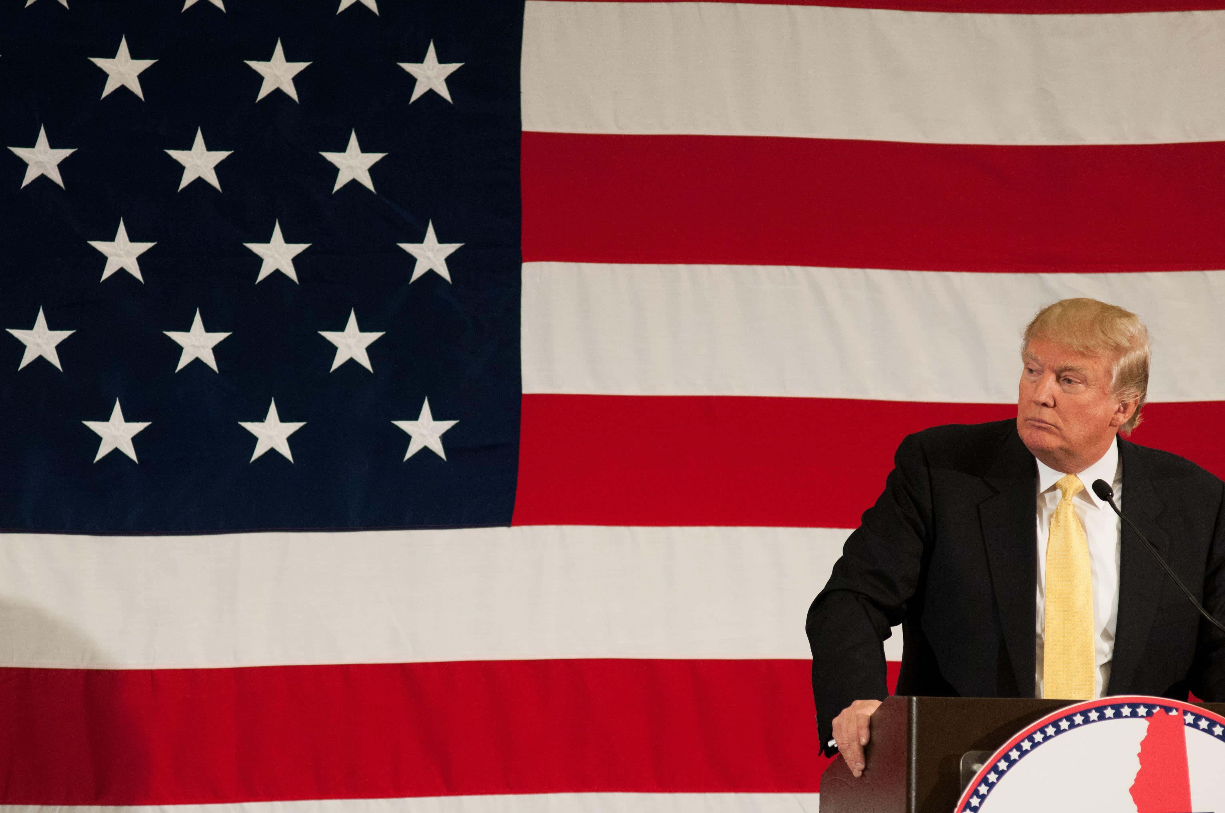 trump 桌布 壁紙 — 當勞·特朗普站在一個講台前，背後有一面大型美國國旗，在正式場合發表演說。