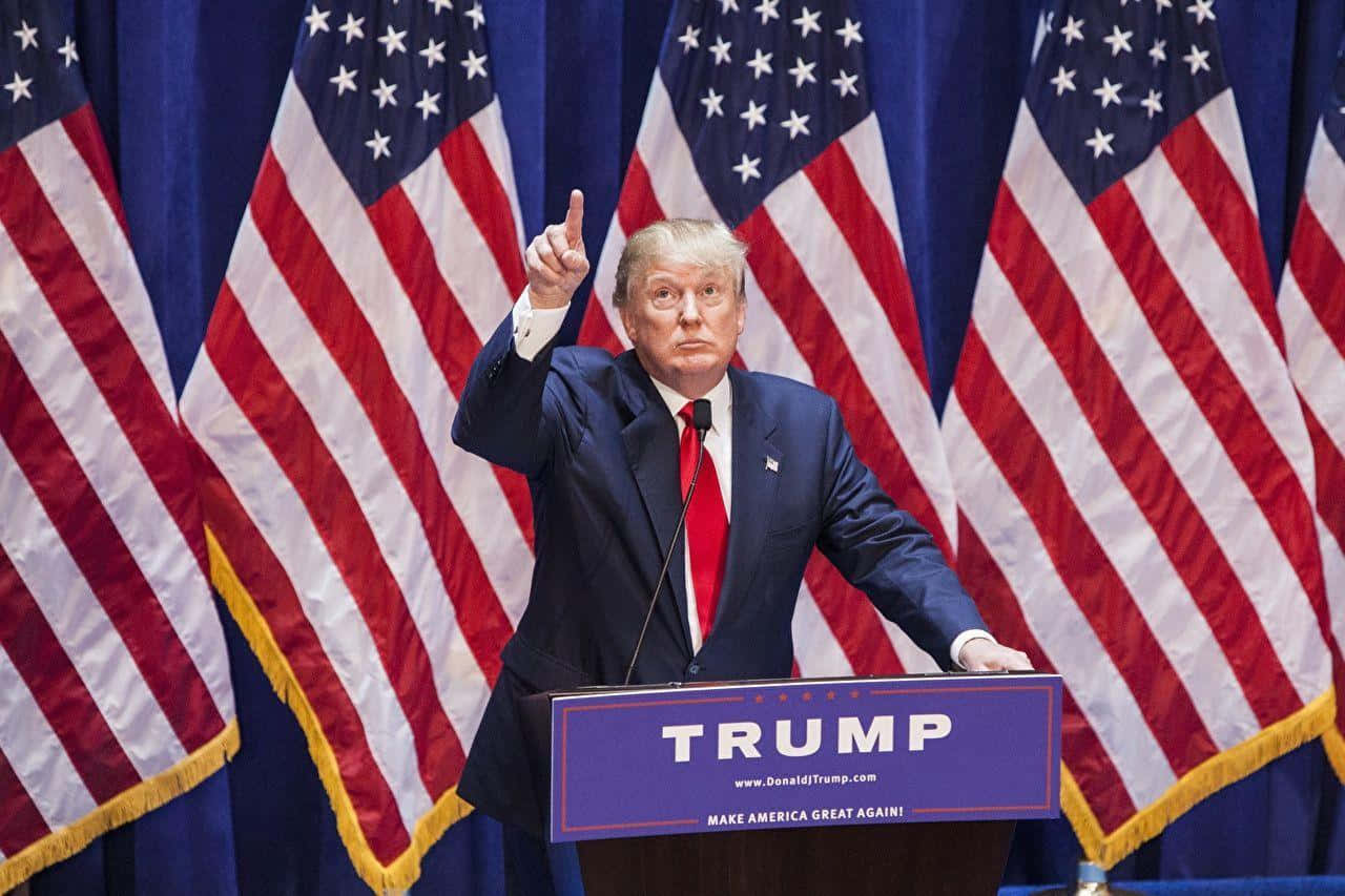 trump 桌布 壁紙 — 當勞·特朗普站在講台上，帶有「TRUMP」和「MAKE AMERICA GREAT AGAIN」標牌，右手食指向上指，背景為多面美國國旗。