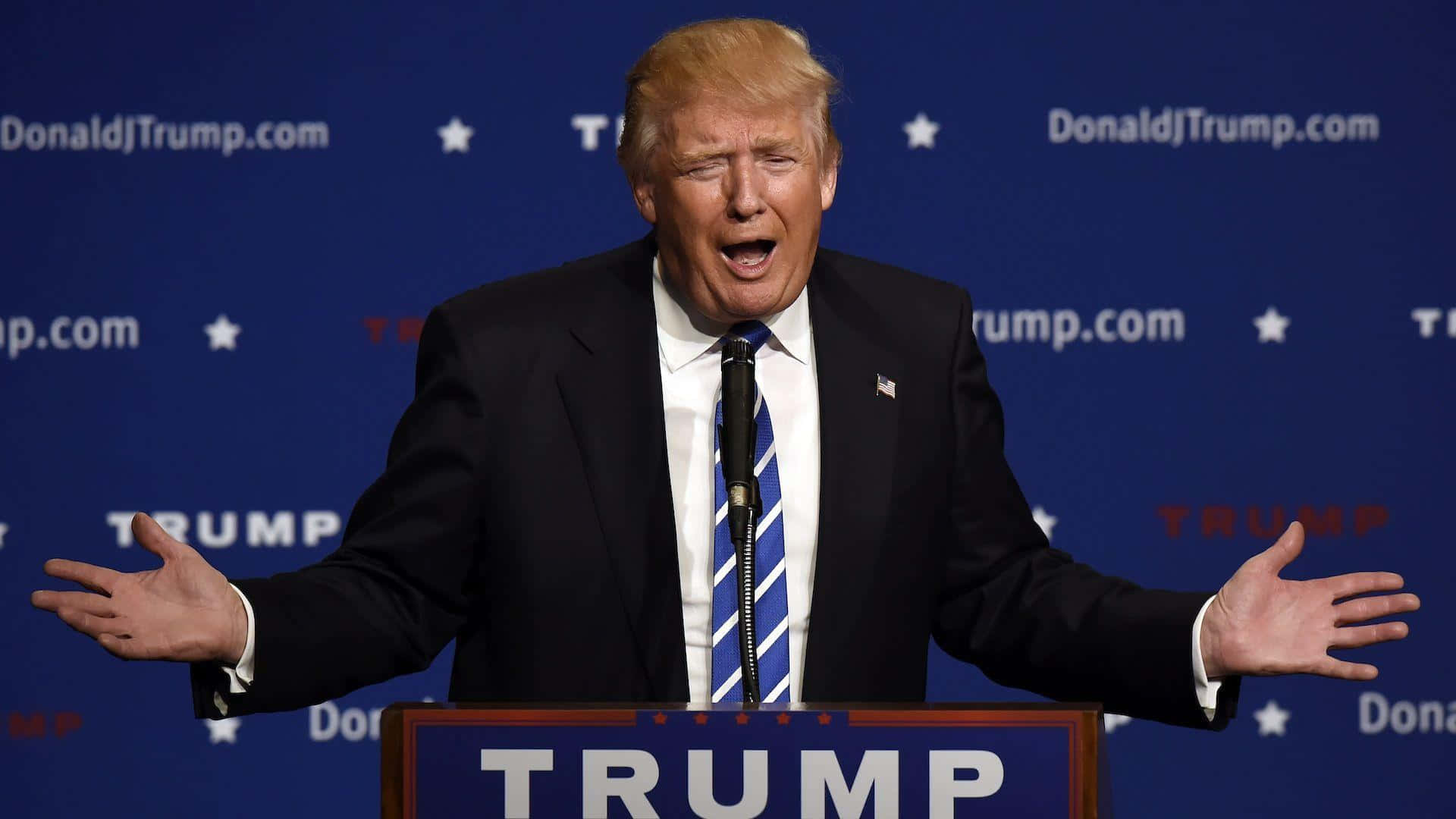 trump 桌布 壁紙 — 當勞·特朗普站在講台上，雙臂張開，在競選活動中大喊，背景為藍色，帶有重複的標誌。
