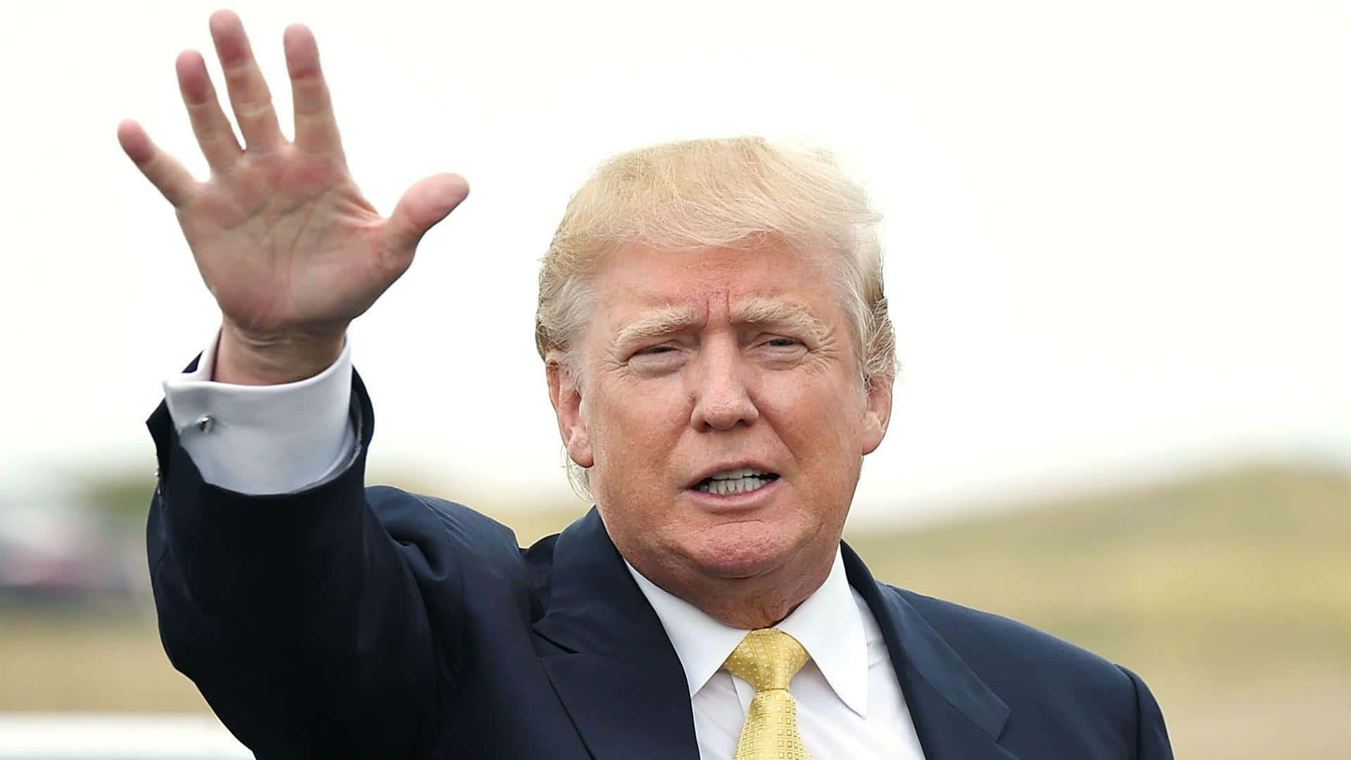 trump 桌布 壁紙 — 當勞·特朗普舉起右手揮手，身穿深色西裝和金色領帶，背景是模糊的戶外環境。