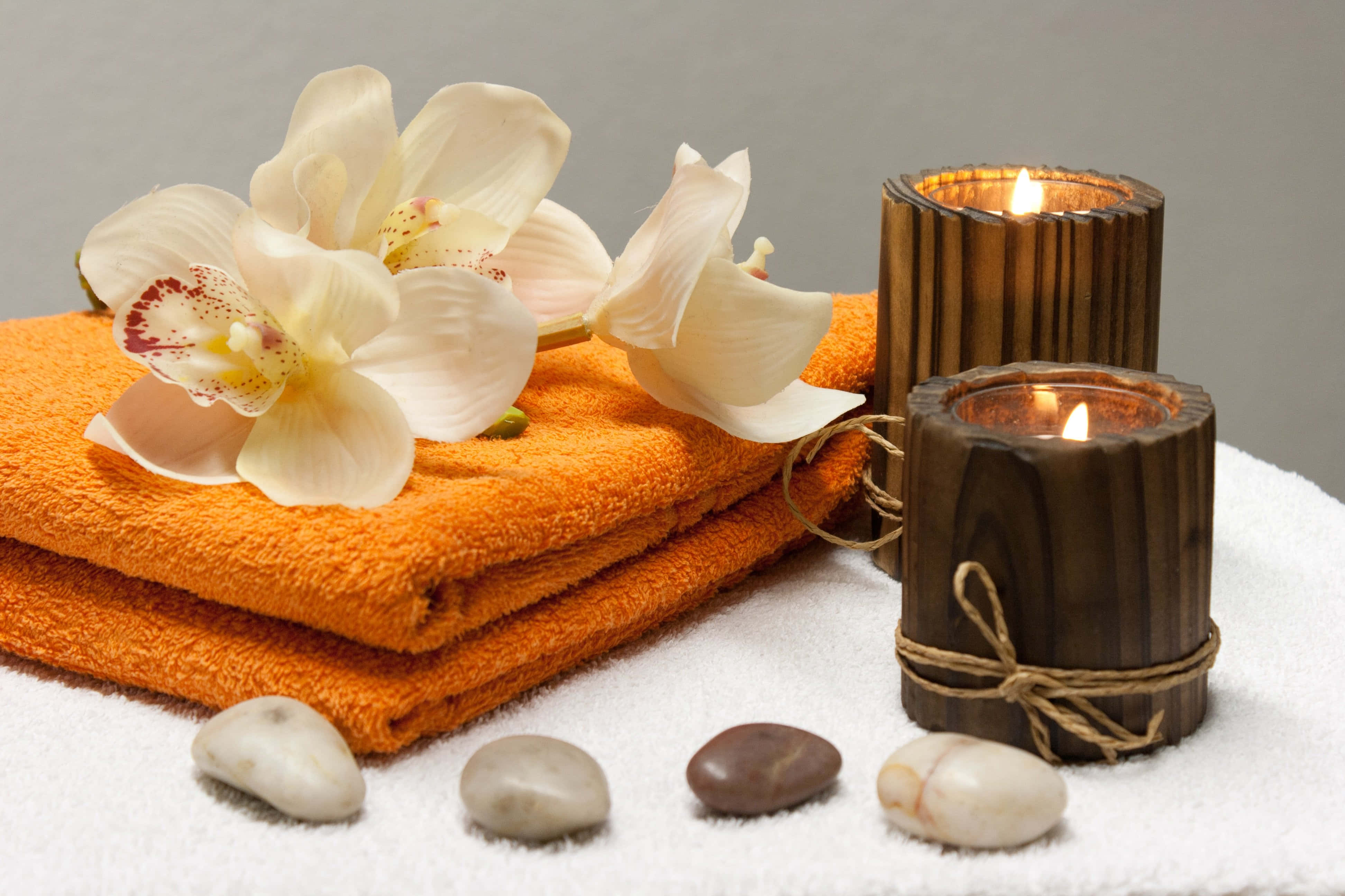 Aromatherapy 桌布 壁紙 — 疊起的橙色毛巾，上面放有白色蘭花，兩側擺放點燃的木蠟燭和光滑的河石。
