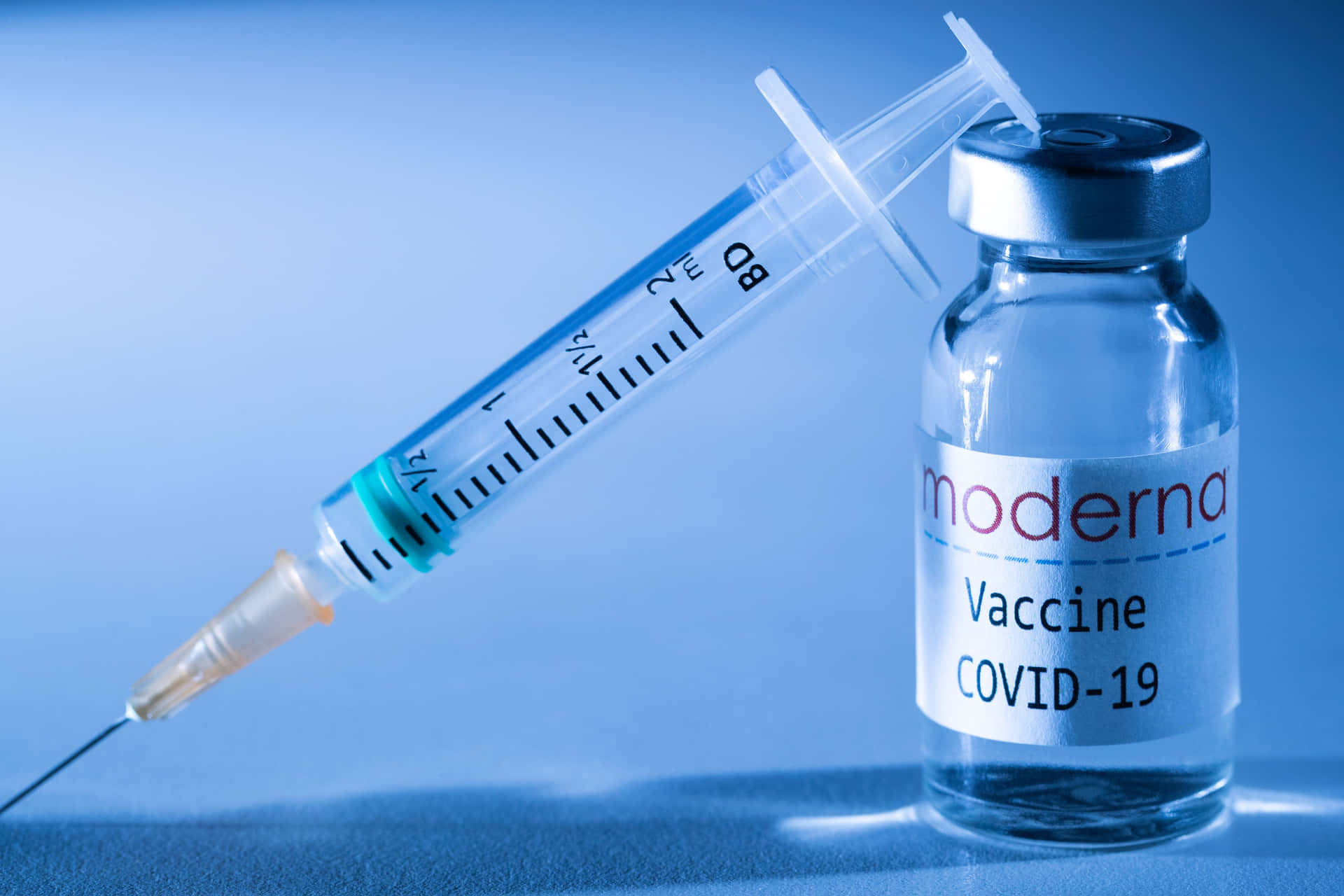 一個Moderna COVID-19疫苗瓶，旁邊有一支注射器，放在藍色表面上。