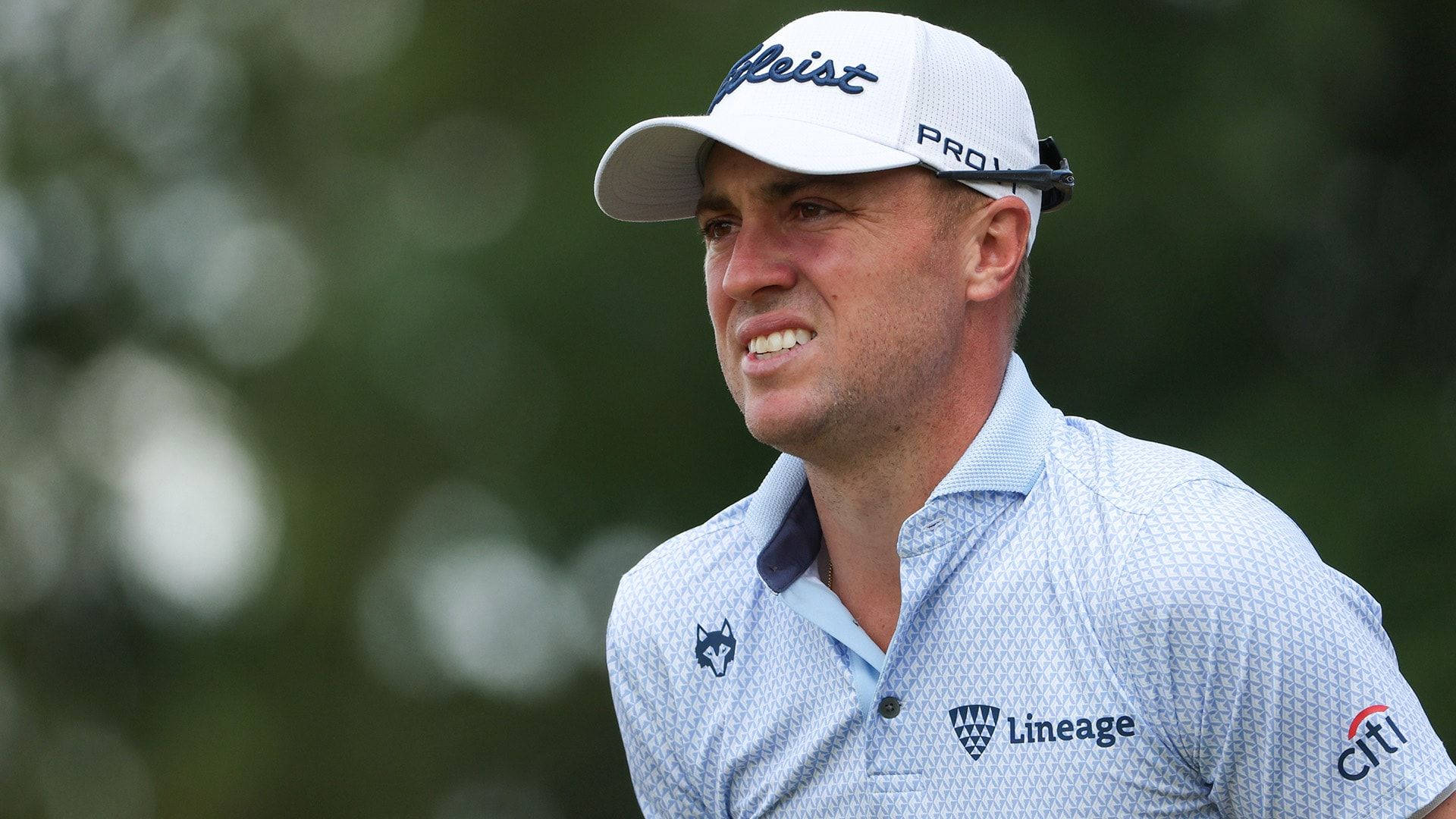 疲憊的 Justin Thomas 壁紙 — 一位專注的高爾夫球手戴著白色帽子，穿著圖案 Polo 衫，在模糊的綠色背景下微微一笑。