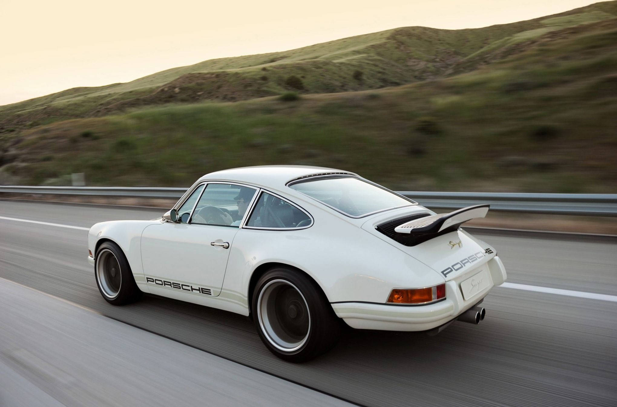 疾馳的Singer Porsche 911 壁紙 — 一輛白色保時捷911在蜿蜒的高速公路上疾馳，背景是金色時刻的起伏綠色山丘。