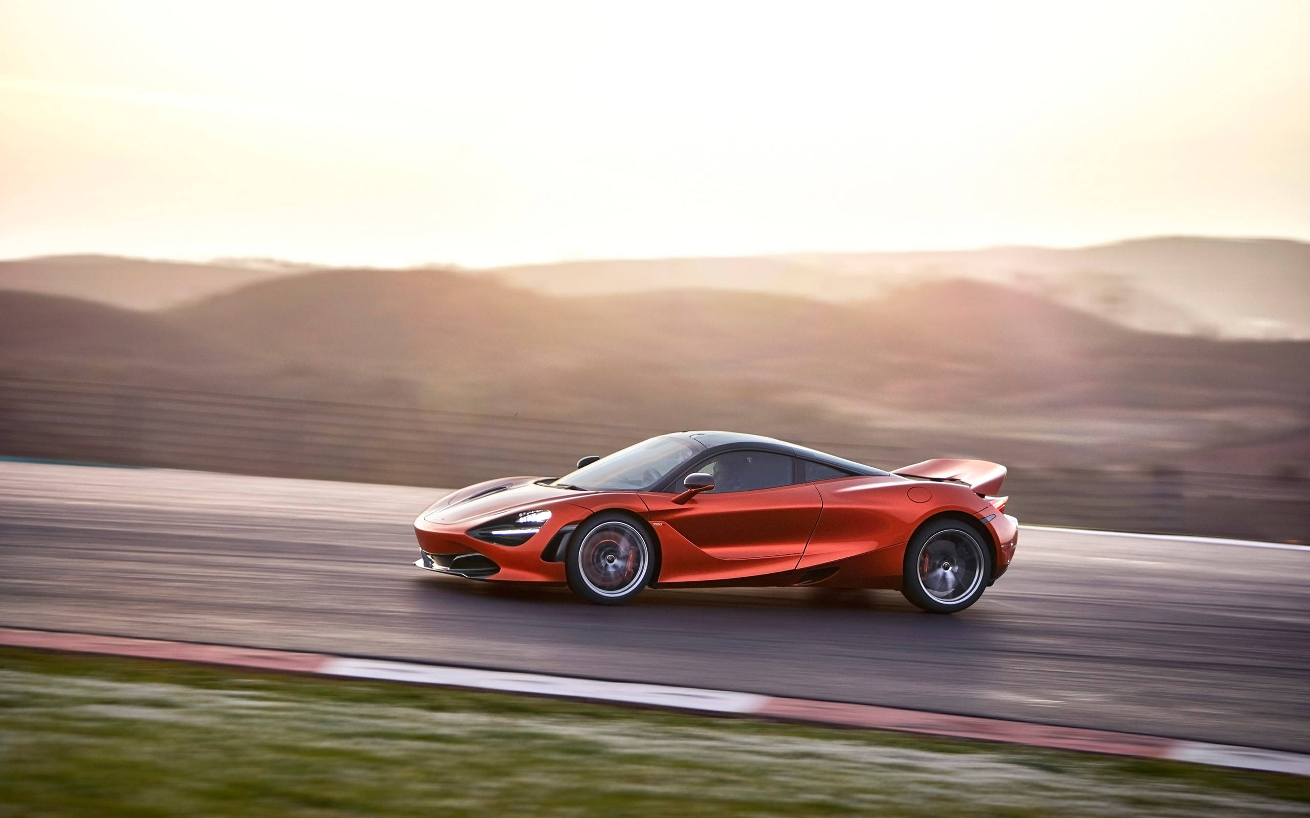疾馳麥拉倫720s 4K 壁紙 — 一輛充滿活力的橙色McLaren 720S在日落時分沿著賽道飛馳，背景是模糊的山丘。
