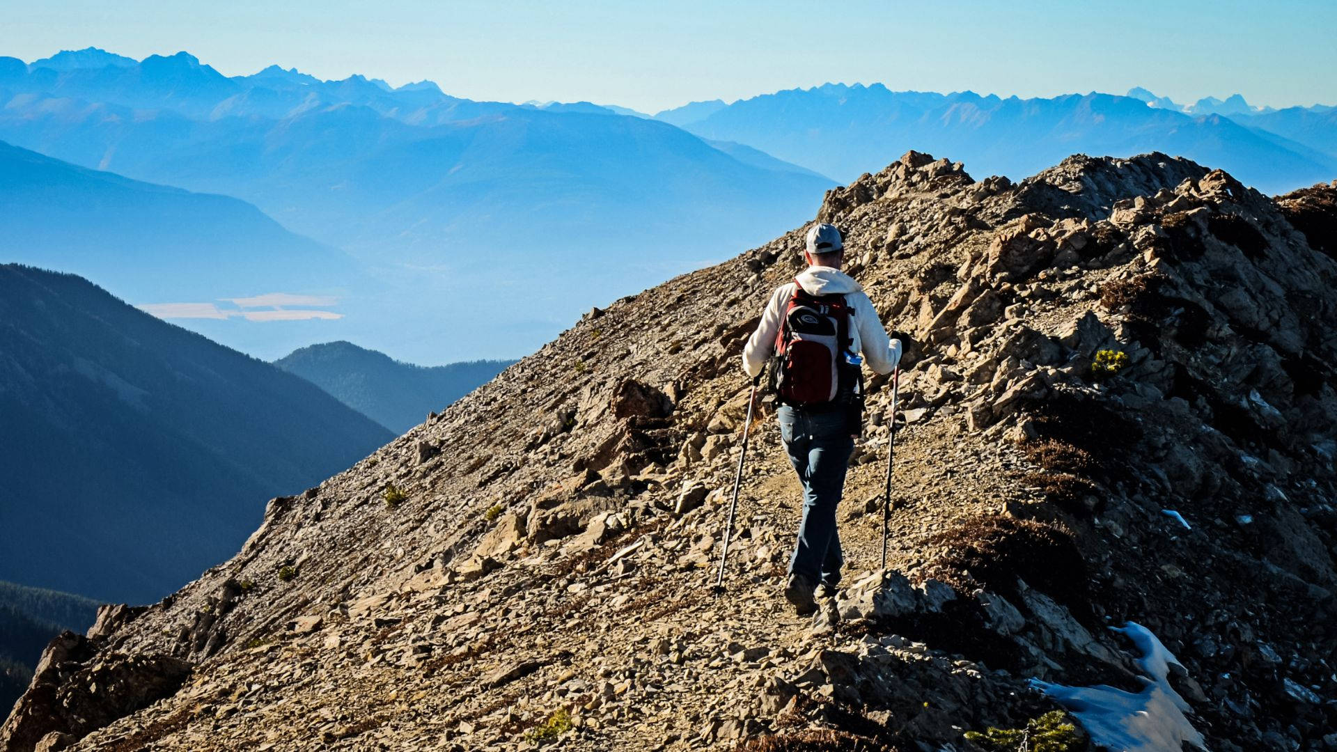 登山客崎嶇山徑之旅 壁紙 — 一位揹著背包、拿著行山杖的遠足者，在晴朗藍天下攀爬岩石山脊，下方遠處可見山峰和山谷。