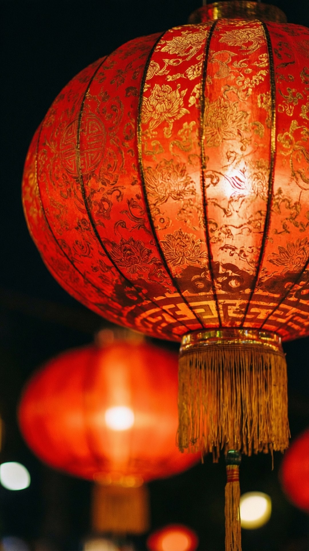 發光燈籠近攝 壁紙 — 一個發光的紅色中國燈籠，帶有金色花卉圖案和流蘇，懸掛在黑暗的夜空背景下，背景有模糊的燈光。