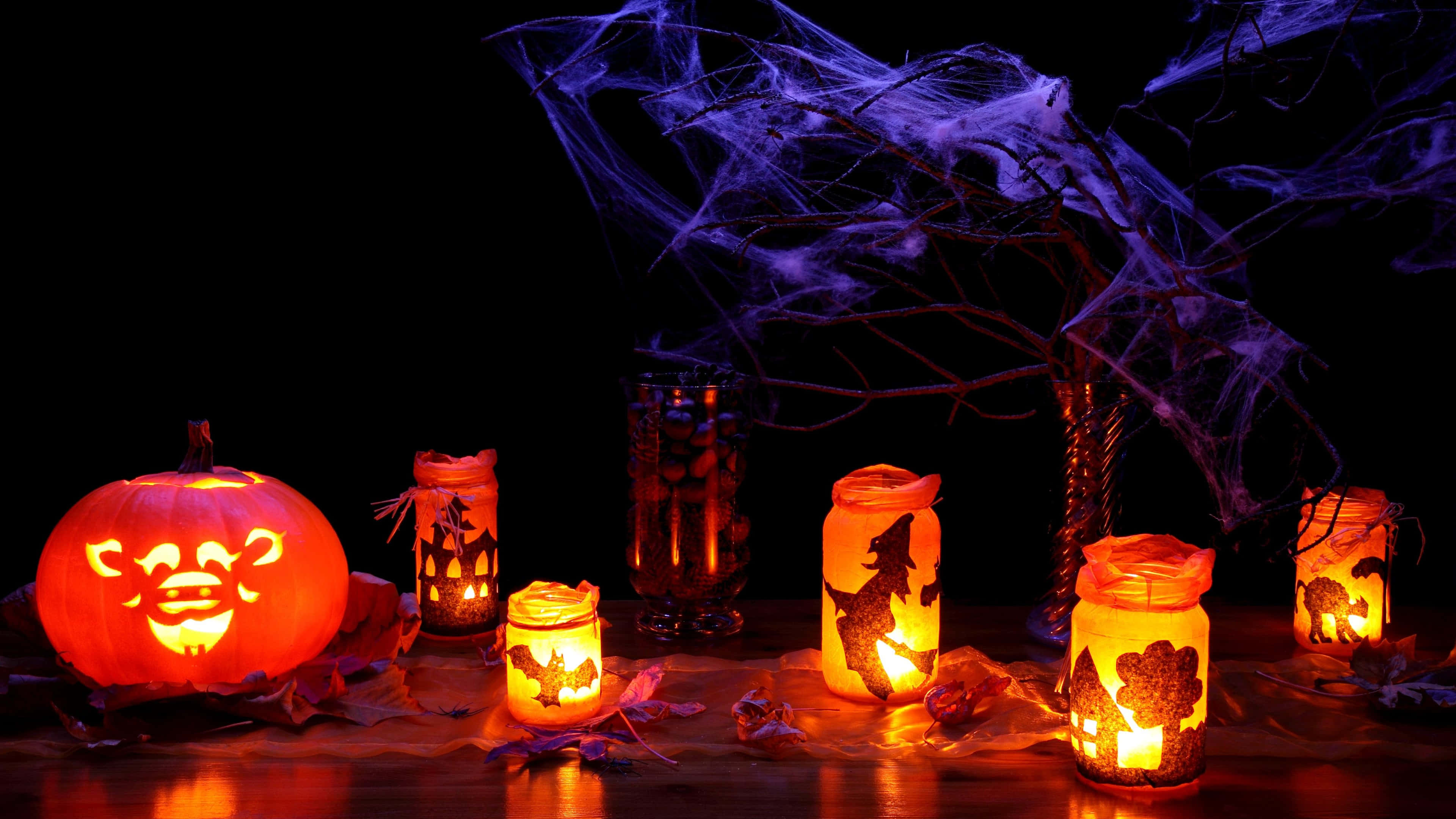 Halloween Candles 桌布 壁紙 — 發光的傑克南瓜燈和女巫主題的罐裝蠟燭在紫色蜘蛛網的黑暗背景下散發出溫暖的光芒。