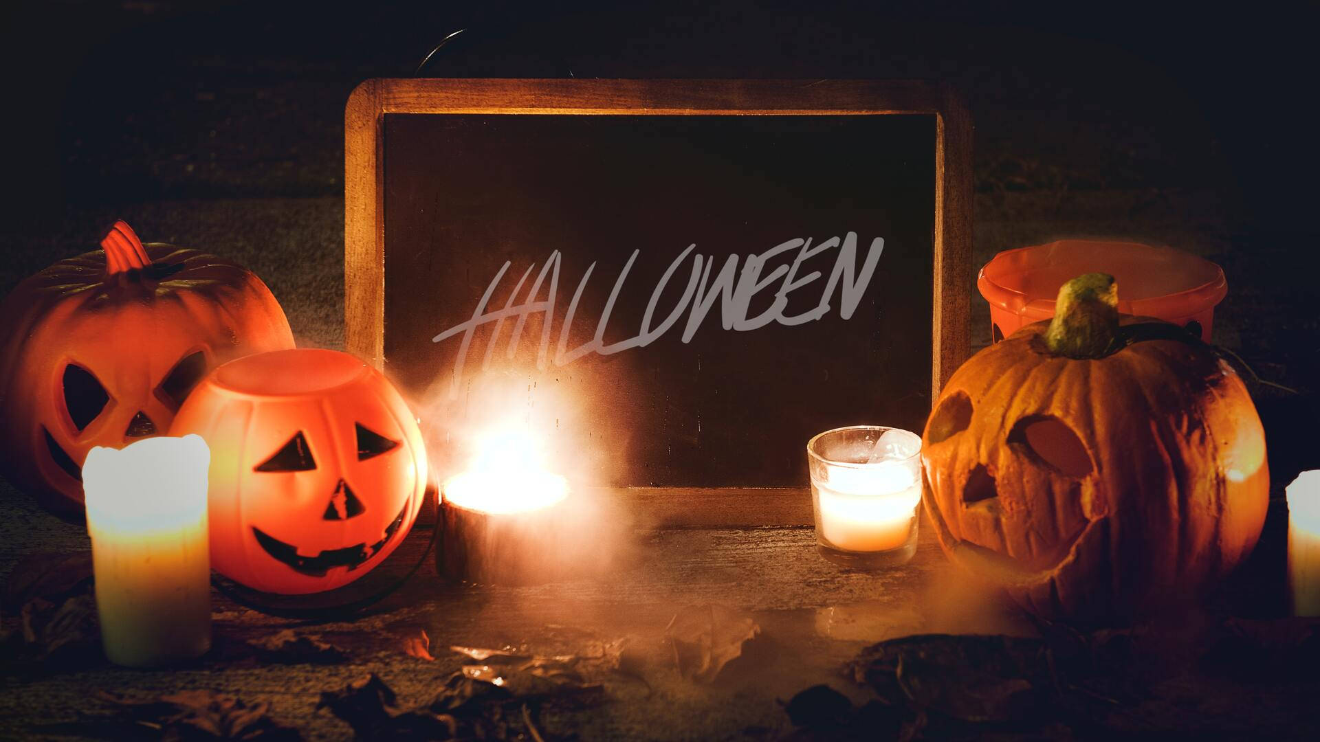 Pumpkins With Candles Halloween Computer 壁紙 — 發光的傑克南瓜燈和搖曳的蠟燭照亮了一個帶有黑板標誌的恐怖萬聖節場景。