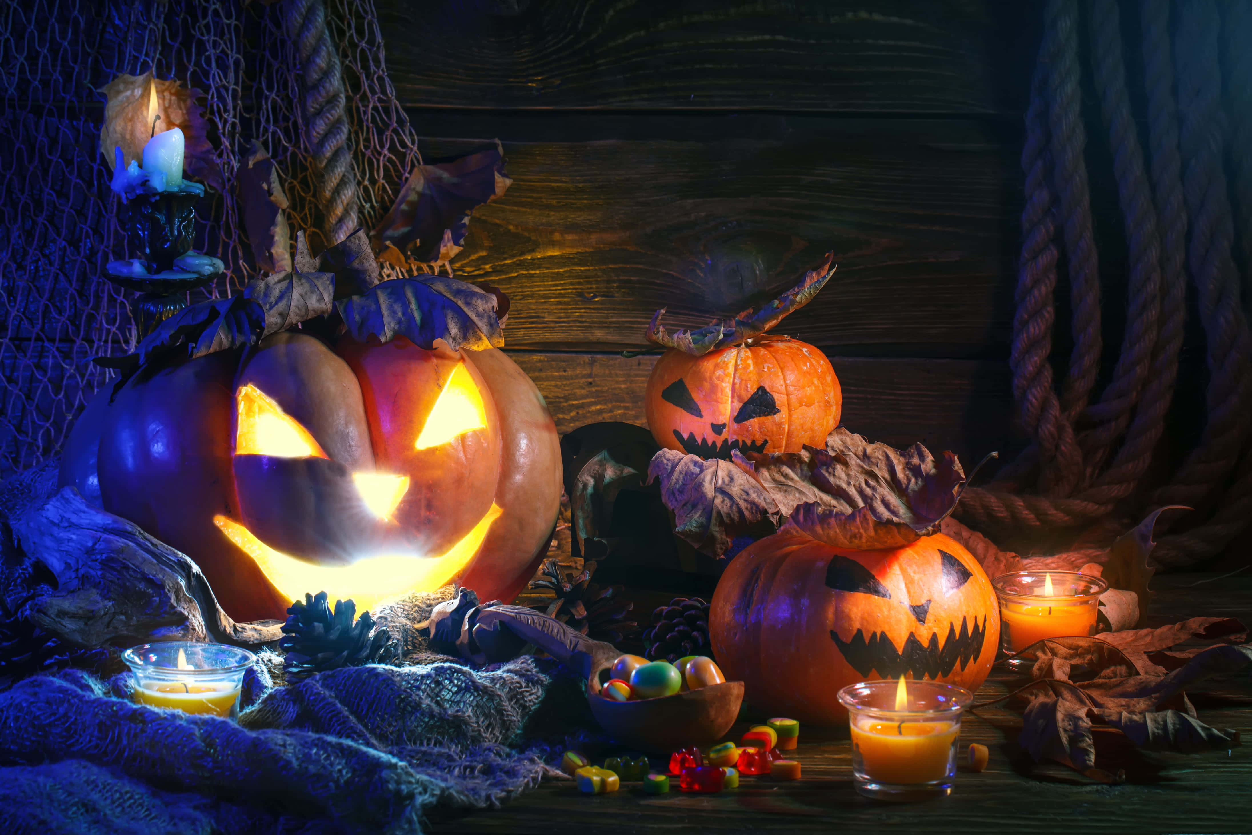 Halloween Candles 桌布 壁紙 — 發光的傑克南瓜燈帶有雕刻面孔，放置在質樸的木桌上，周圍環繞著蠟燭、秋葉和糖果，在昏暗的藍色和橙色燈光下。