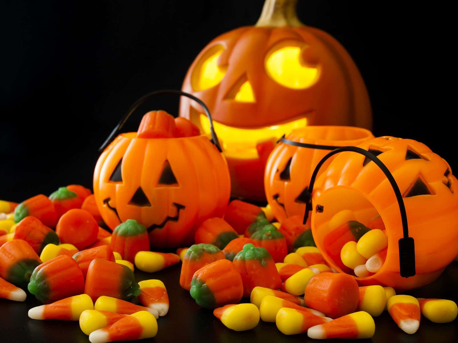 Halloween Treats 桌布 壁紙 — 發光的傑克南瓜燈桶和糖果玉米在溫暖的萬聖節燈光下散落在黑暗的表面上。