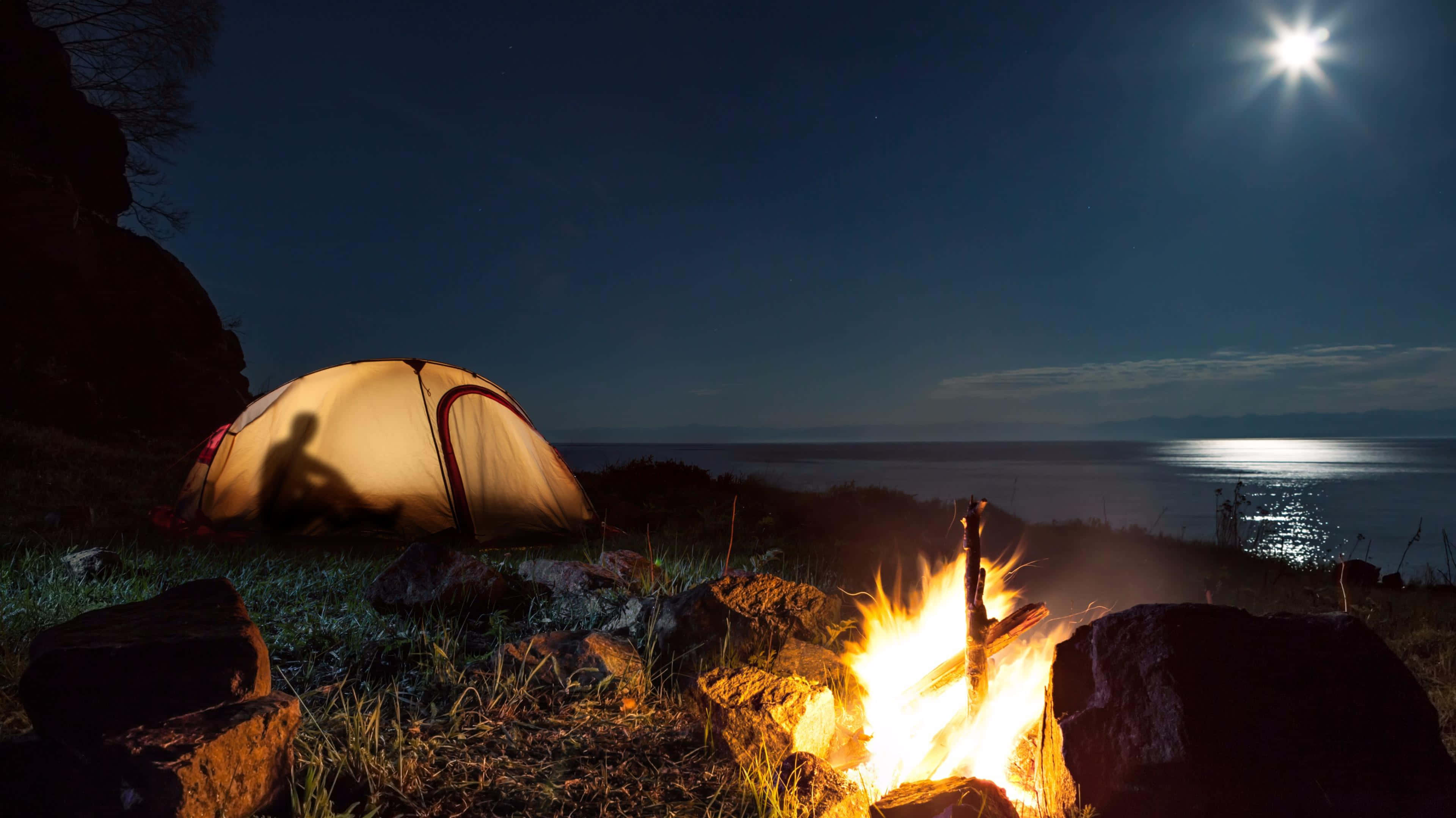 Campfire 桌布 壁紙 — 發光的帳篷和噼啪作響的營火照亮了岩石海岸，上方是平靜水面上的明亮月亮。