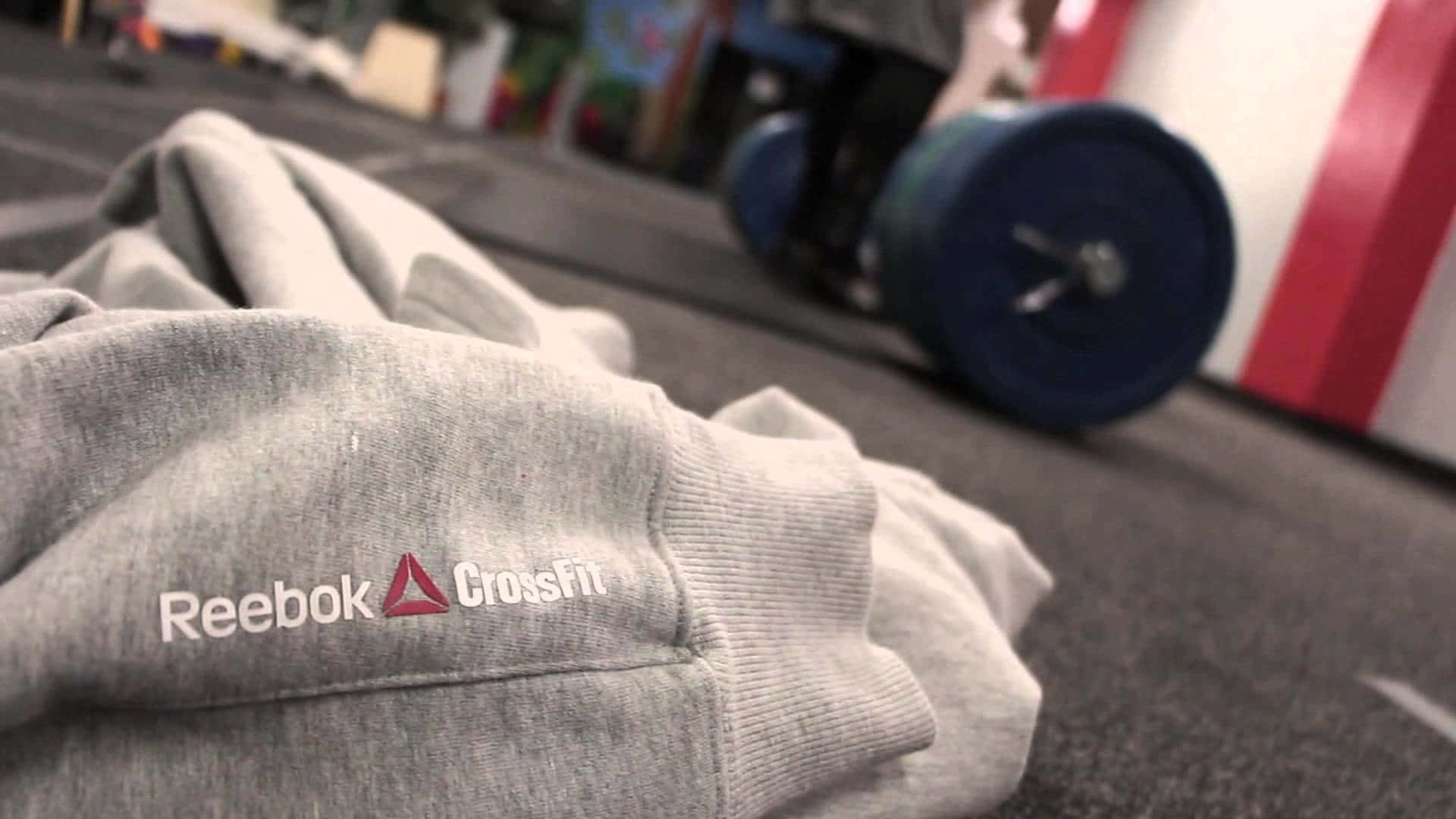 發揮潛能，於Reebok中感受無敵自信 壁紙 — 灰色 Reebok CrossFit 運動衫的特寫，放在健身室地板上，背景有藍色槓鈴。