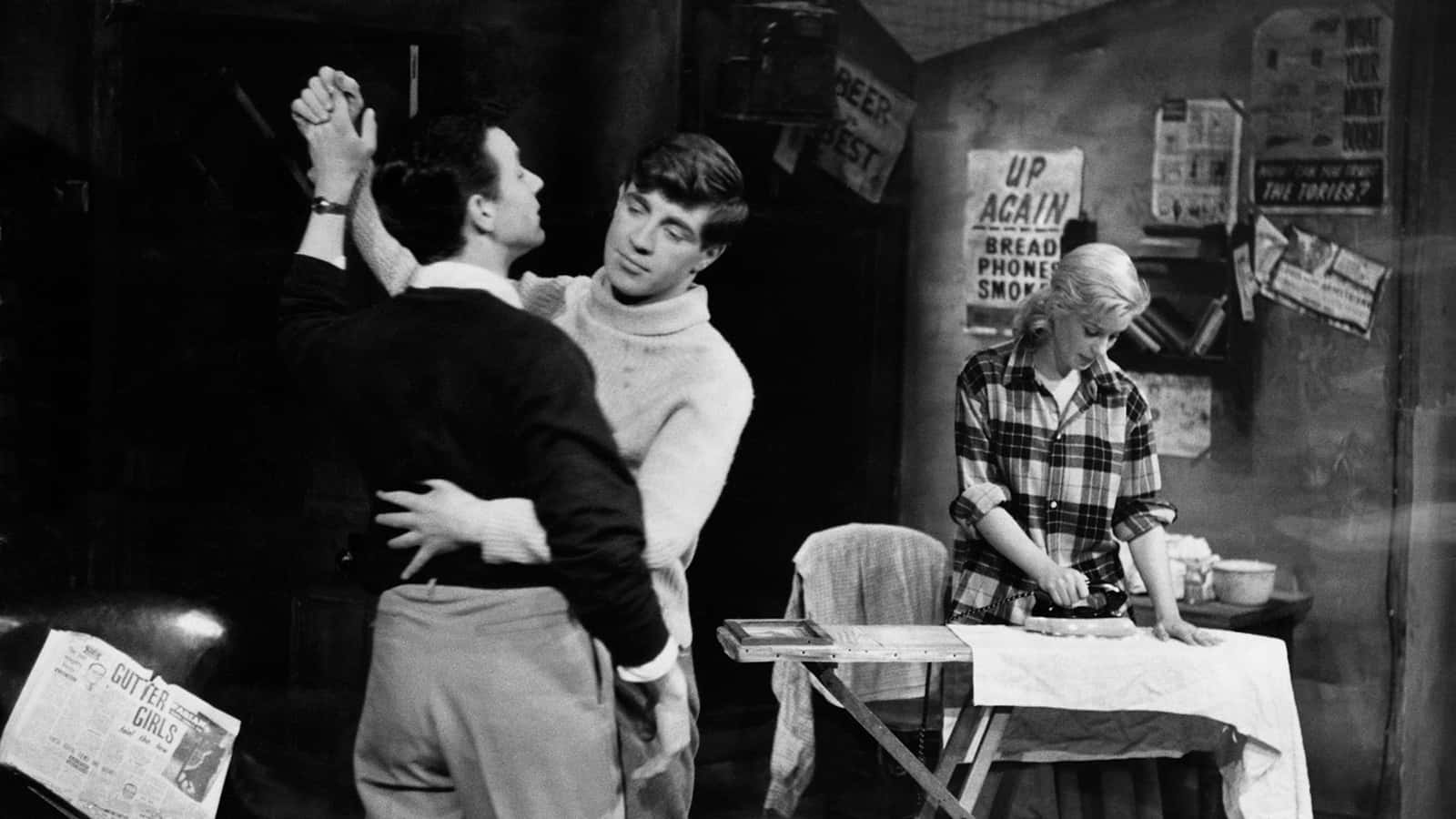 兩個男人在1960年代的酒吧裡擁抱，而一個女人在附近熨衣服。