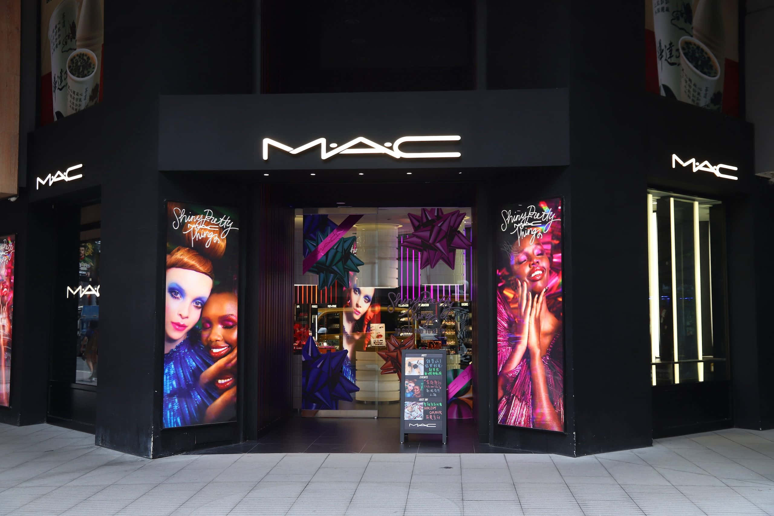 發現反映你風格的彩妝 壁紙 — 一間時尚的黑色MAC化妝品店，帶有發光招牌和充滿活力的紫色、粉紅色和藍色宣傳海報，模特兒妝容大膽。