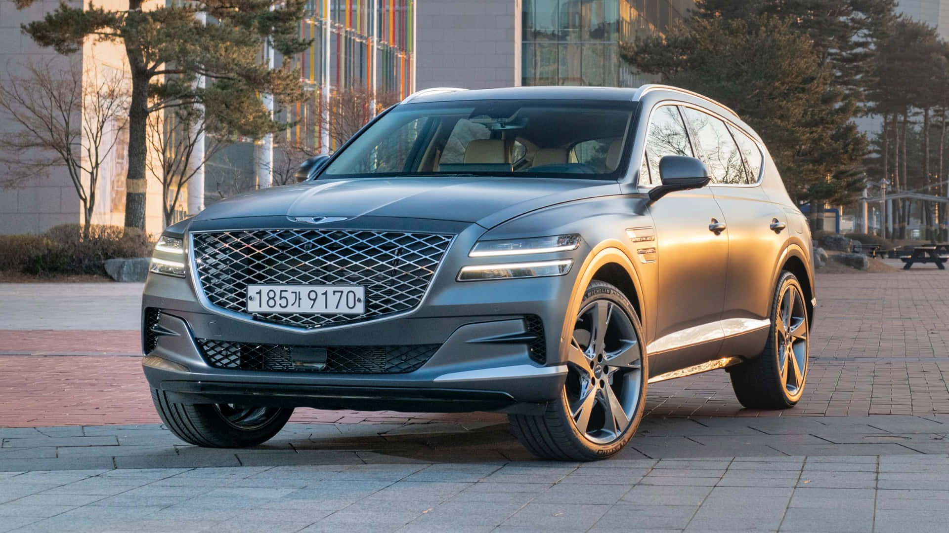 灰色 Genesis GV80 SUV 停泊在鋪砌的廣場上，靠近現代建築和樹木。