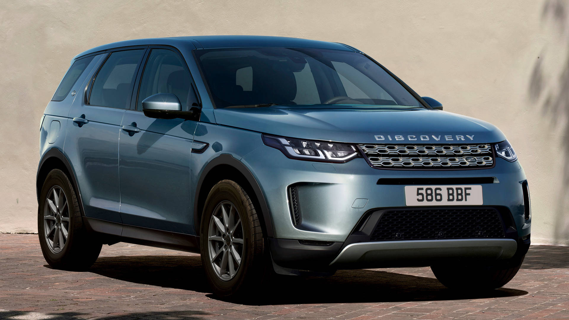 發現運動版路虎 iPhone 主題壁紙 壁紙 — 一輛時尚的藍色 Land Rover Discovery SUV 停泊在磚砌路面上，背景是米色牆壁，在明亮的日光下。
