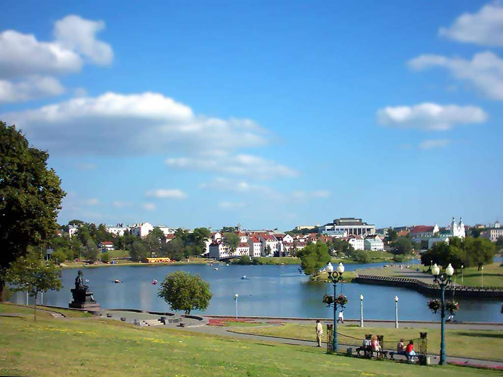 白俄羅斯公園湖泊 壁紙 — 湖邊公園晴朗的一天，人們在長椅上放鬆，船隻在水上，周圍是綠樹和城市建築，藍天白雲。