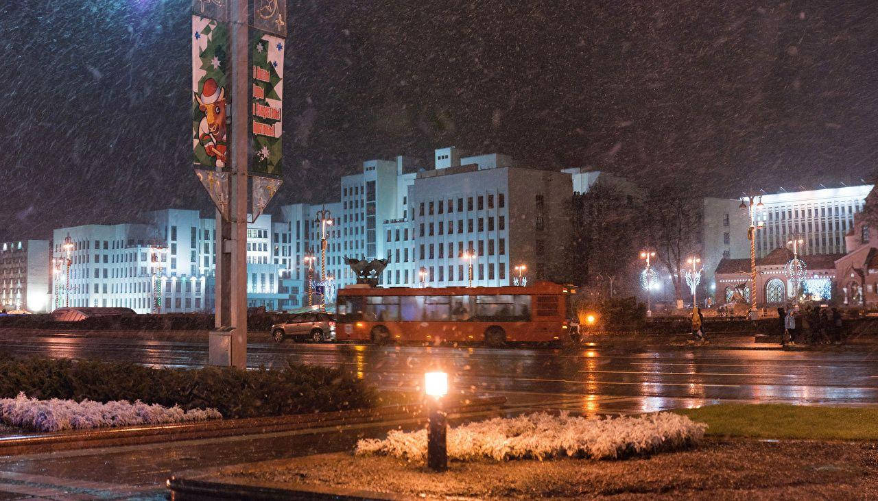 白俄羅斯冬季城市 壁紙 — 夜晚，大雪覆蓋濕漉漉的城市街道，一輛紅色巴士在閃爍的路燈和節日標誌下駛過。