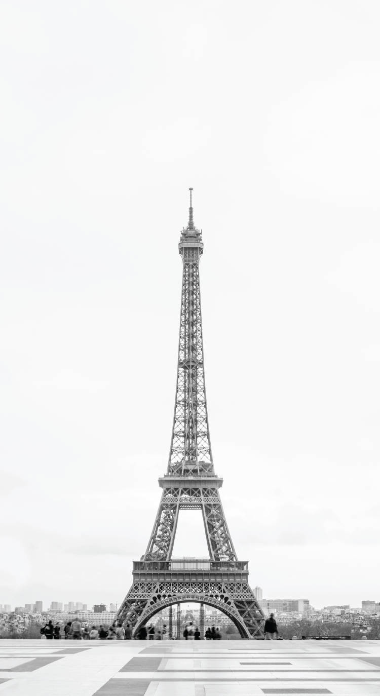 白天空與埃菲爾鐵塔 iPhone 壁紙 — 艾菲爾鐵塔在陰雲密佈的天空下高聳入雲的黑白景觀，塔底聚集著人群。