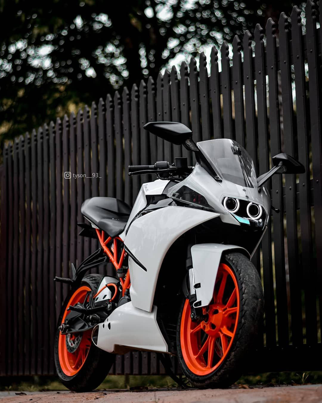 白橙配色 KTM RC 200 壁紙 — 一架時尚的白色和橙色運動電單車，帶有黑色點綴，停在深色木柵欄旁，光線柔和。