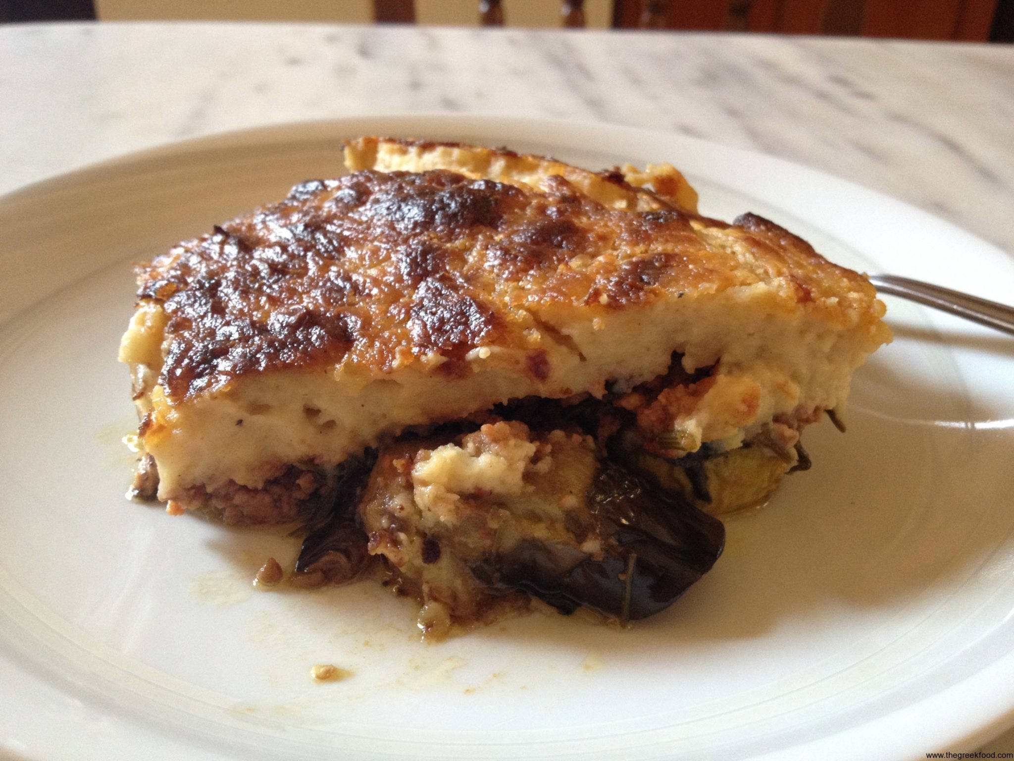 白瓷盤上的穆薩卡切片 壁紙 — 一塊金黃色的千層茄子（Moussaka），表面酥脆，可見茄子層，放在白色碟上。