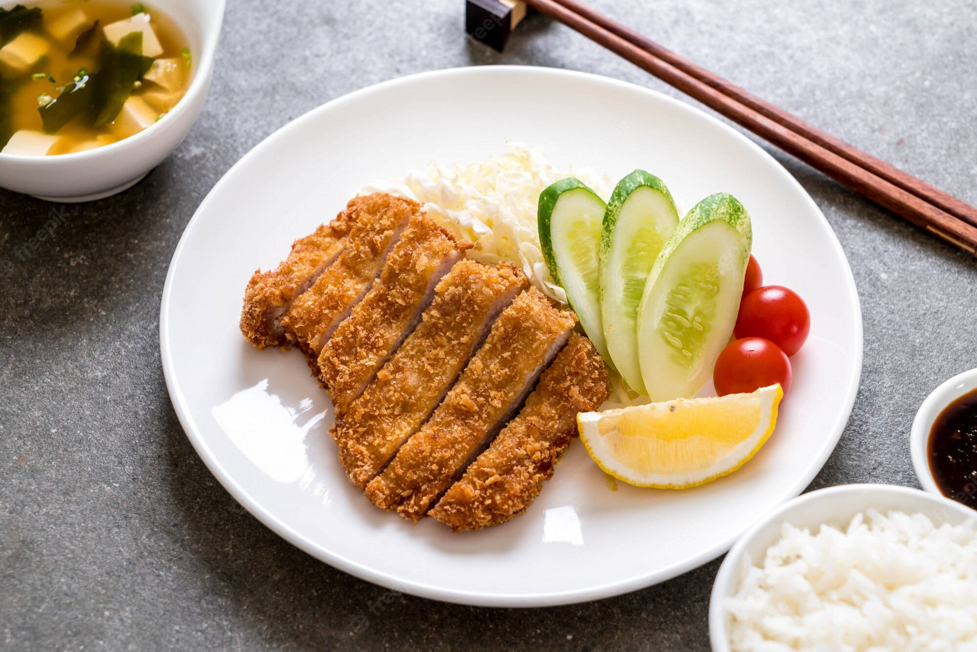 白盤上的炸豬排 壁紙 — 金黃色切好的炸豬排，配上米飯、青瓜、番茄、檸檬和味噌湯，放在灰色桌上。