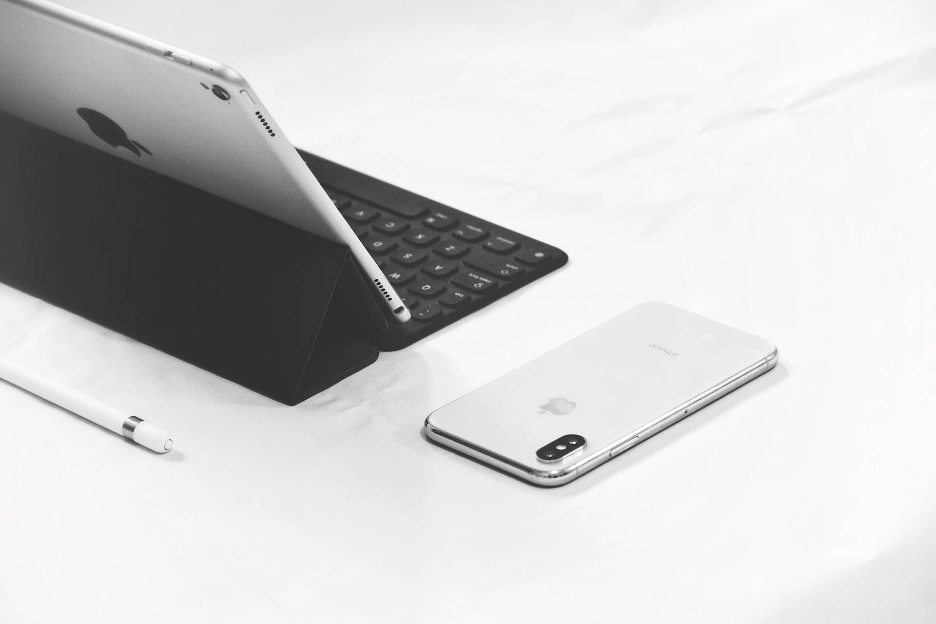 銀色 iPad 配備鍵盤保護套和 iPhone 在白色表面上，附有 Apple Pencil。
