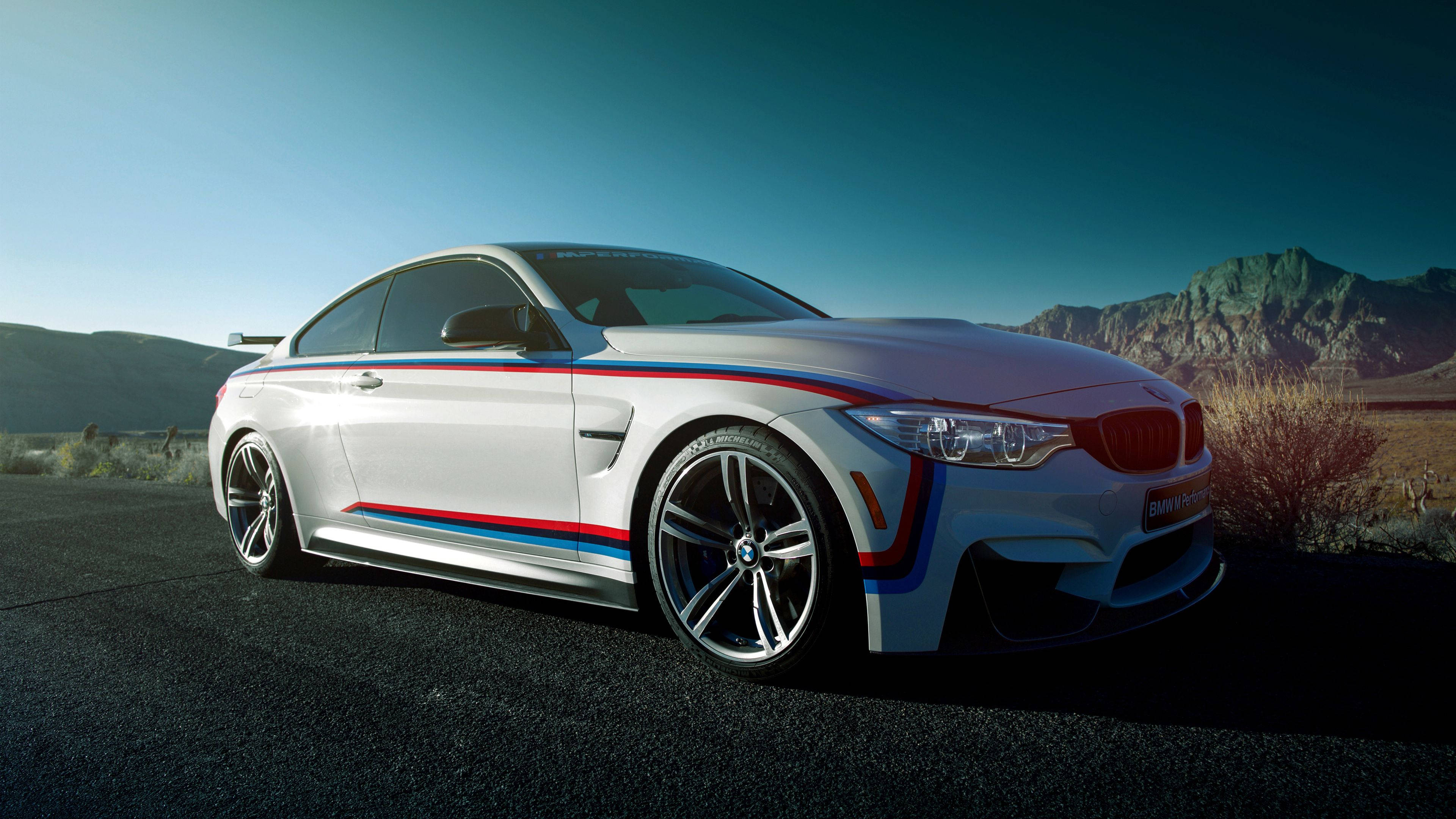 白色 BMW M 系列 壁紙 — 一輛白色BMW M4雙門跑車，帶有紅色、藍色和黑色賽車條紋，停在沙漠公路上，背景是崎嶇的山脈和晴朗的藍天。