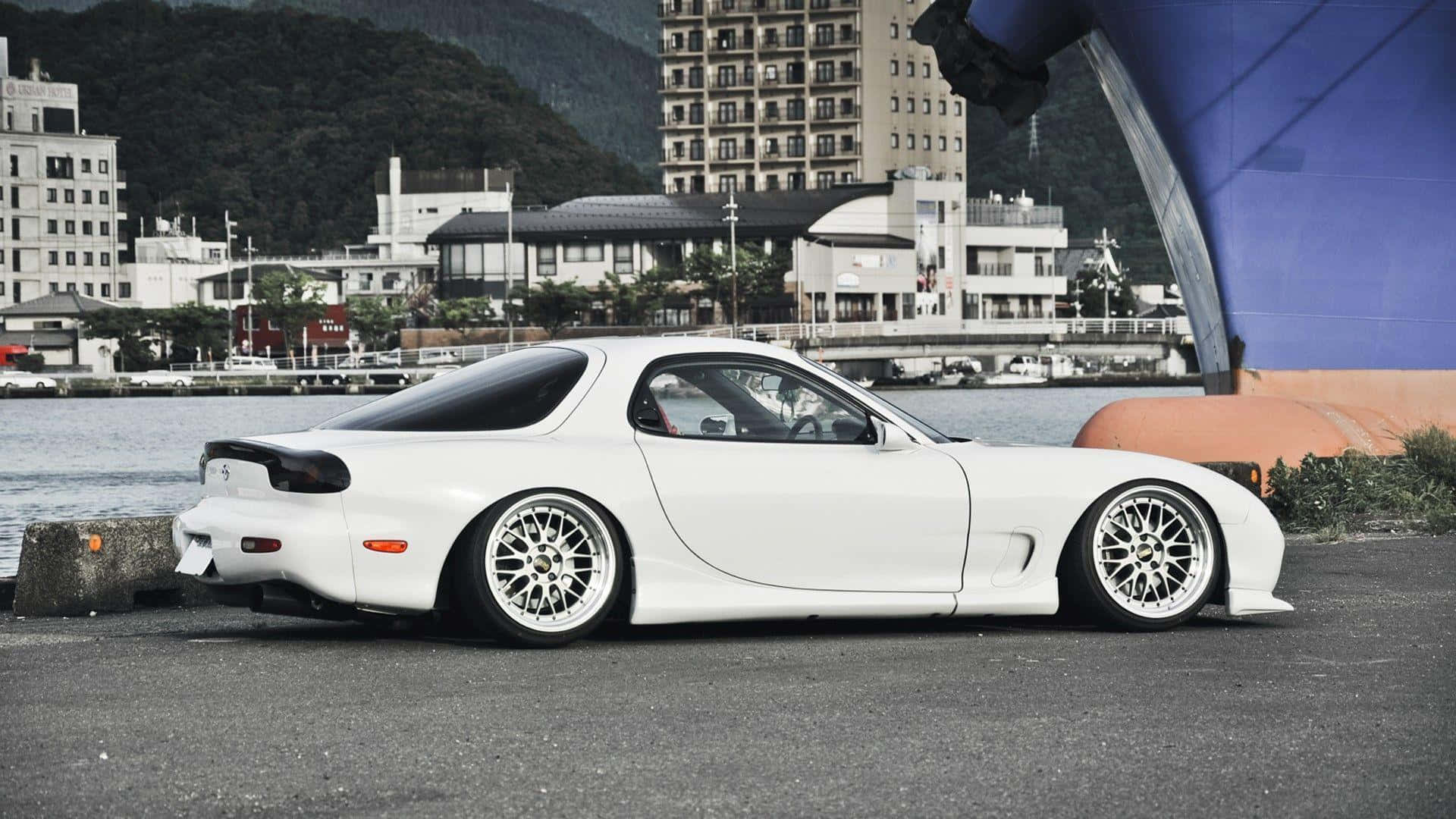白色萬事得RX-7跑車停泊在水邊，背景有城市建築物。