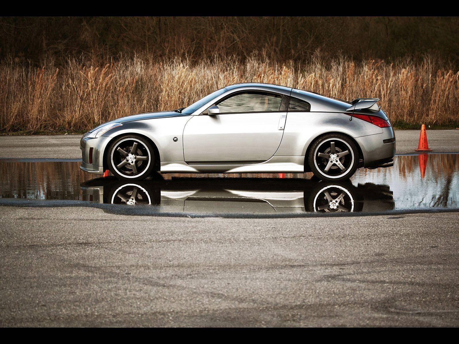白色 Nissan 350z 壁紙 — 一輛銀色跑車完美地倒映在濕瀝青賽道的水坑中，周圍是乾燥的蘆葦。