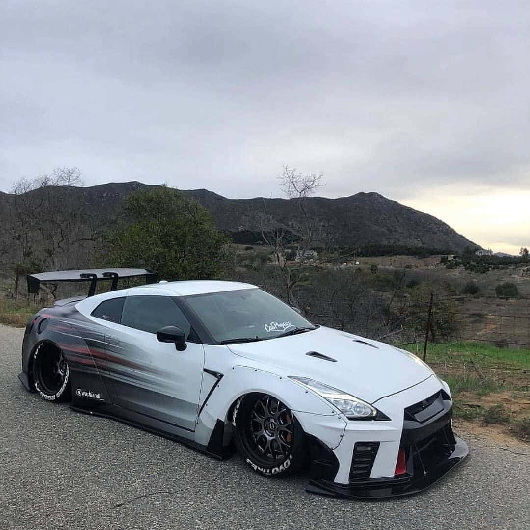 白色 Nissan R35 GT-R 山景圖 壁紙 — 一架白色和黑色改裝的日產GT-R跑車停泊在碎石路上，背景是多雲的山脈和稀疏的樹木。