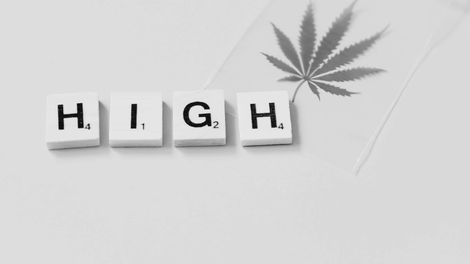 Black And White Word 桌布 壁紙 — 白色 Scrabble 字母磚拼出 HIGH，旁邊有一片大麻葉，置於簡約灰色表面上。