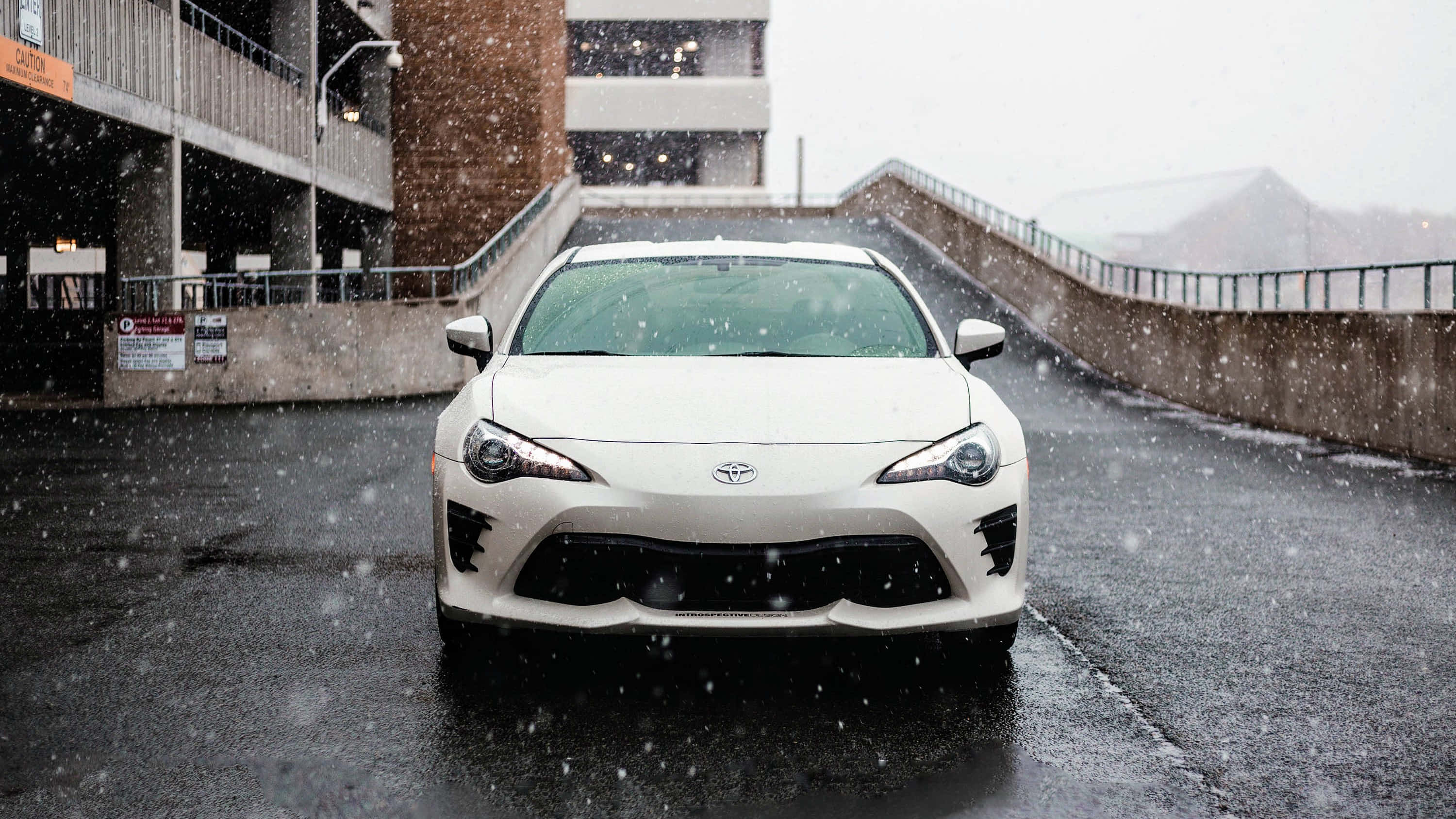 白色 Toyota Supra 停泊於風景如畫的背景 壁紙 — 一輛白色跑車在積雪的城市道路上向前行駛，背景有飄落的雪花和混凝土結構。