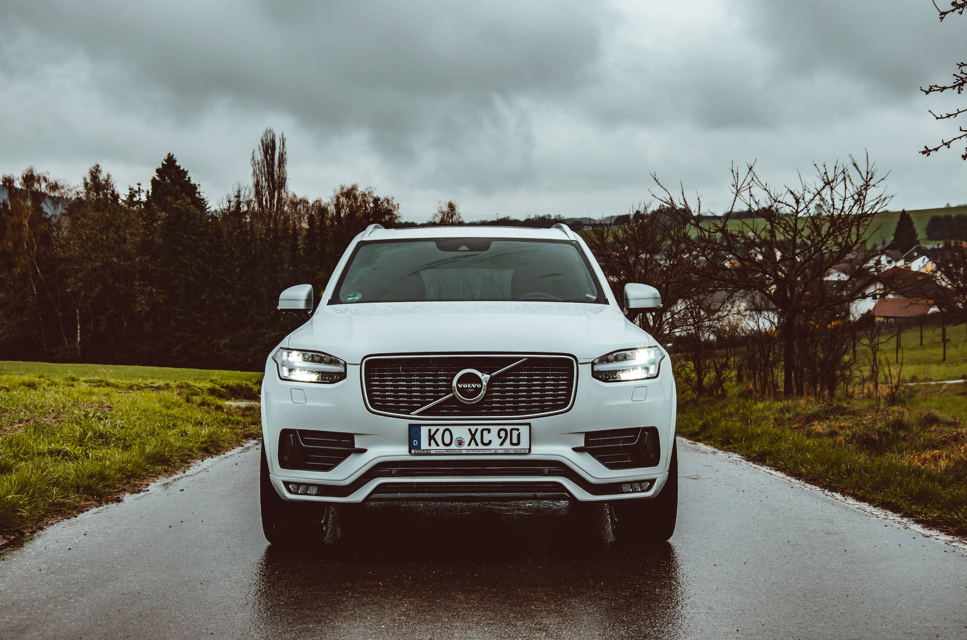白色Volvo XC90 SUV在潮濕的鄉村道路上，多雲天空和綠色田野。