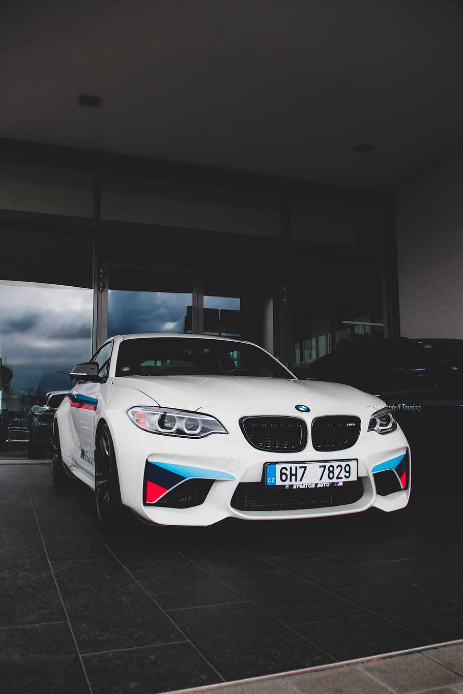 白色BMW M2跑車，帶有藍色和紅色點綴，停放在室內靠近玻璃門處，顯示多雲天空。