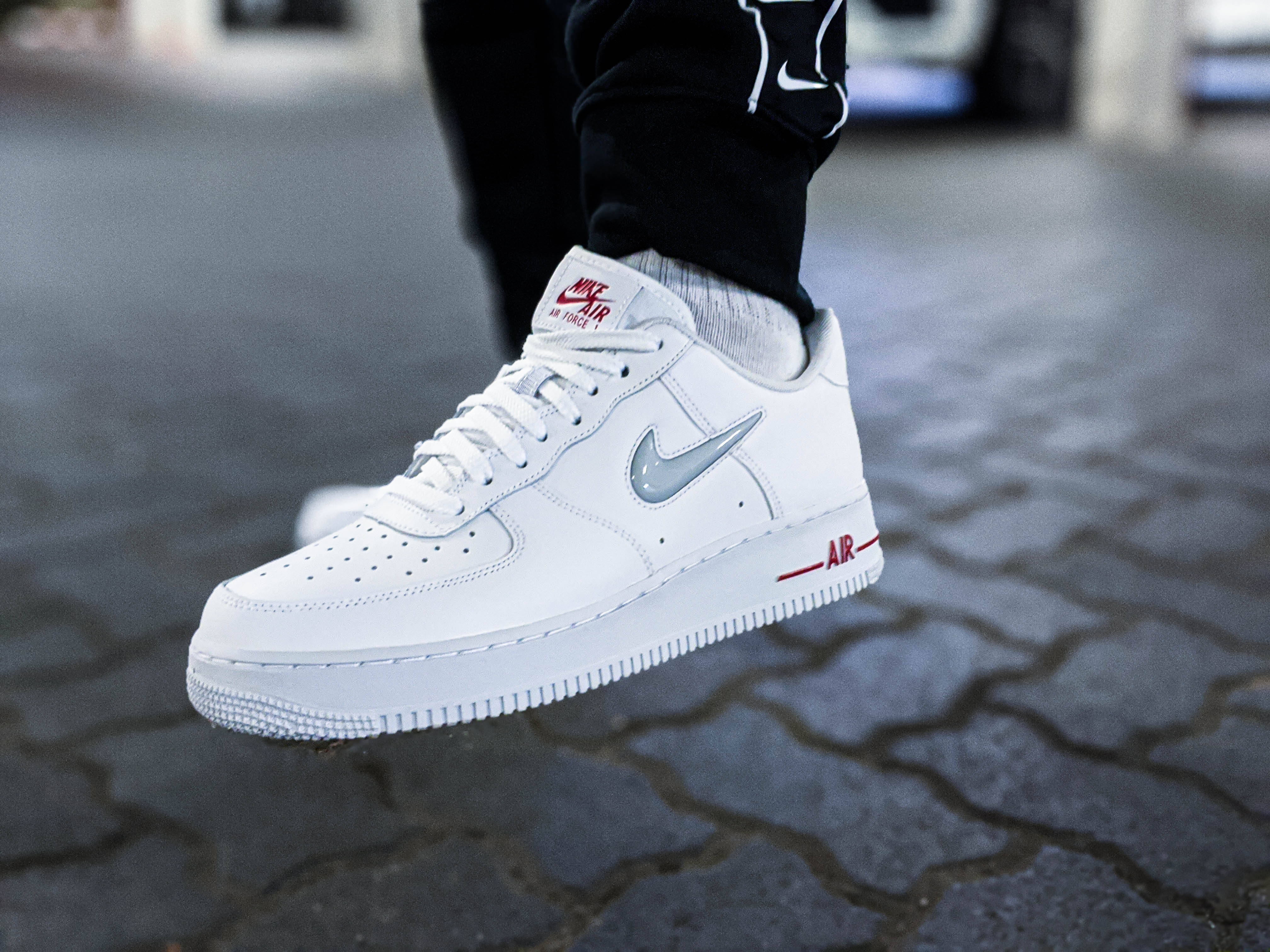 白色Nike Air橡膠運動鞋 壁紙 — 白色Nike Air Force 1運動鞋特寫，帶有紅色和銀色點綴，放在鵝卵石街道上。